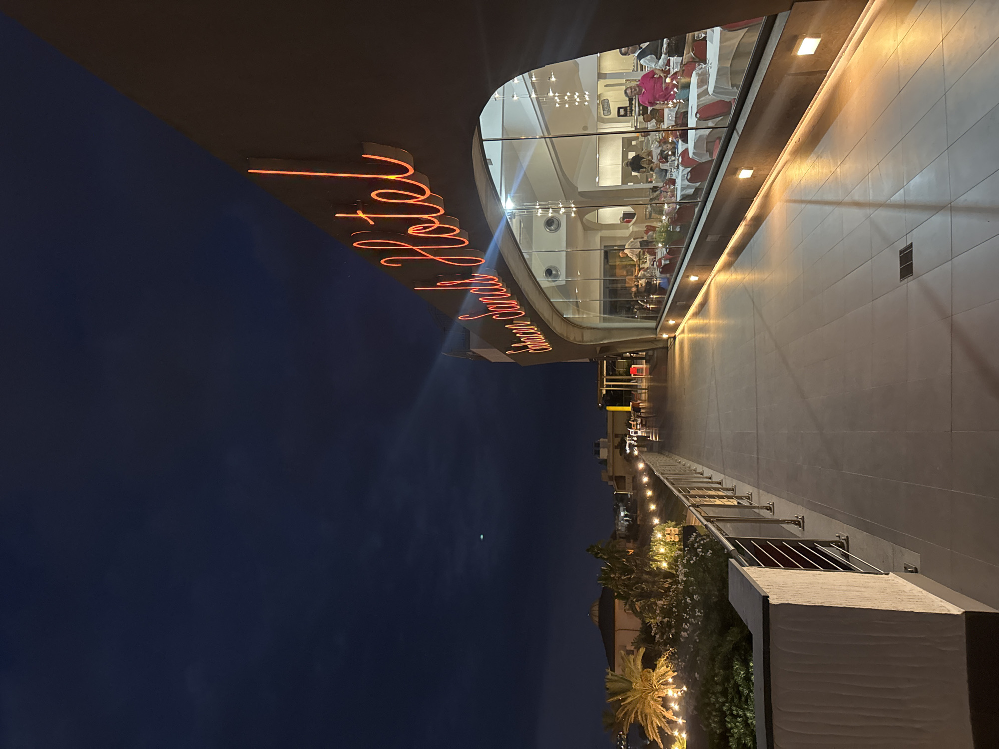 In the city of Capaci, Italy, an outdoor perspective view captures a building at twilight, displaying a neon sign reading "Saracen Sands Hotel" in a cursive font. The building's facade curves inward, featuring a large glass window that overlooks an interior restaurant. Inside, patrons are seated at tables adorned with white tablecloths and red chairs. Waitstaff can be seen attending to customers. The exterior walkway, tiled and illuminated by strategically placed square lights, extends to the left, bordered by a low white wall and a metal railing. Beyond the railing, palm trees and lush greenery are visible, along with a string of decorative lights creating a festive atmosphere. The sky above is a deep blue, transitioning into night, with a faint celestial object visible. The overall scene suggests a pleasant evening at a seaside establishment.