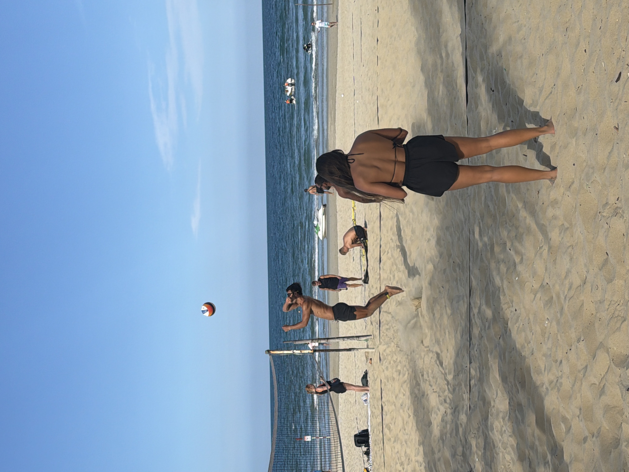 This image depicts a vibrant beach scene in Đà Nẵng, Vietnam, under a bright, clear blue sky. The sandy beach fills the foreground and right side, meeting the blue-green ocean that extends into the distance on the left.
The central activity is a game of beach volleyball. A man, wearing dark shorts and sunglasses, is captured in mid-air, having jumped to spike a colorful volleyball (red, white, and blue/black) over a dark net. His body is nearly horizontal to the ground as he extends his arm towards the ball. Another player is visible near the net on the opposite side.
Beyond the volleyball court, several other people are present. A woman with long dark hair, wearing a black bikini top and shorts, stands with her back to the viewer, gazing towards the ocean. Further out in the water, multiple jet skis or similar watercraft are visible with people onboard.
The bright sunlight casts distinct shadows on the sand, indicating a clear, sunny day, likely during the late morning or early afternoon. The scene conveys an atmosphere of active recreation and leisure at the beach. No visible text is present in the image.