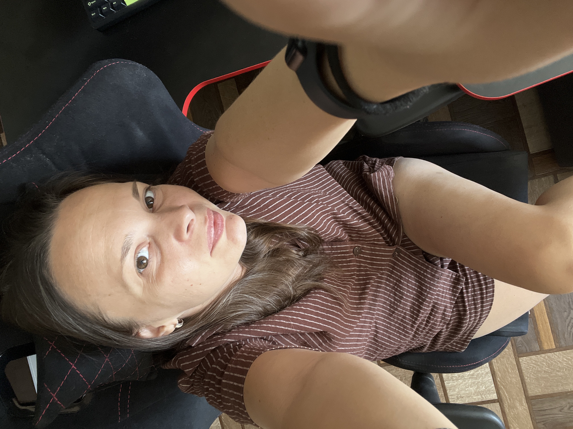 A woman is sitting in a black gaming chair with red trim. She is wearing a brown and white striped shirt. Her left arm is extended towards the camera, holding a phone to take a selfie. The chair is on a light brown wooden floor. A black keyboard is partially visible to the right of the chair. The image is taken from a high angle, looking down at the woman. There is no visible indication of the city of Tolyatti, Russia, in the image.