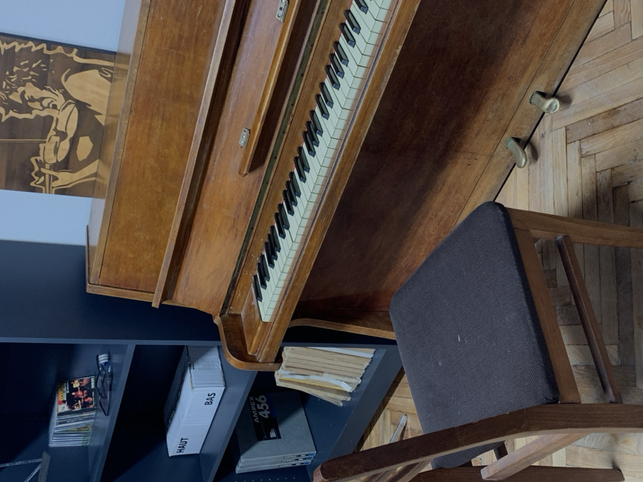 The image displays an indoor scene in the city of Kyiv, Ukraine. A vintage-style, wooden upright piano, dark amber in color, occupies the right and center of the frame. Its white and black keys show minor wear and dust. Two brass foot pedals are visible at the piano's base. To the left, a dark grey multi-shelf unit is positioned. The top shelf holds a stack of compact discs, a pair of dark-framed glasses, and a small black electronic device. The middle shelf contains a white cardboard box labeled "HAUT" and "BAS." Below this, several grey items marked "AMPEX 456" are stored. A stack of light-colored papers or thin books rests against the lower shelf and the side of the piano. In the foreground, a wooden chair with a dark fabric-upholstered seat is placed in front of the piano, oriented towards it. The floor throughout the visible area is light brown parquet wood. Mounted on the light blue-grey wall above the piano is a wooden relief depicting a figure with curly hair playing a flute-like instrument. The wall behind the dark grey shelving unit appears to be a darker shade. No individuals are present in the scene.