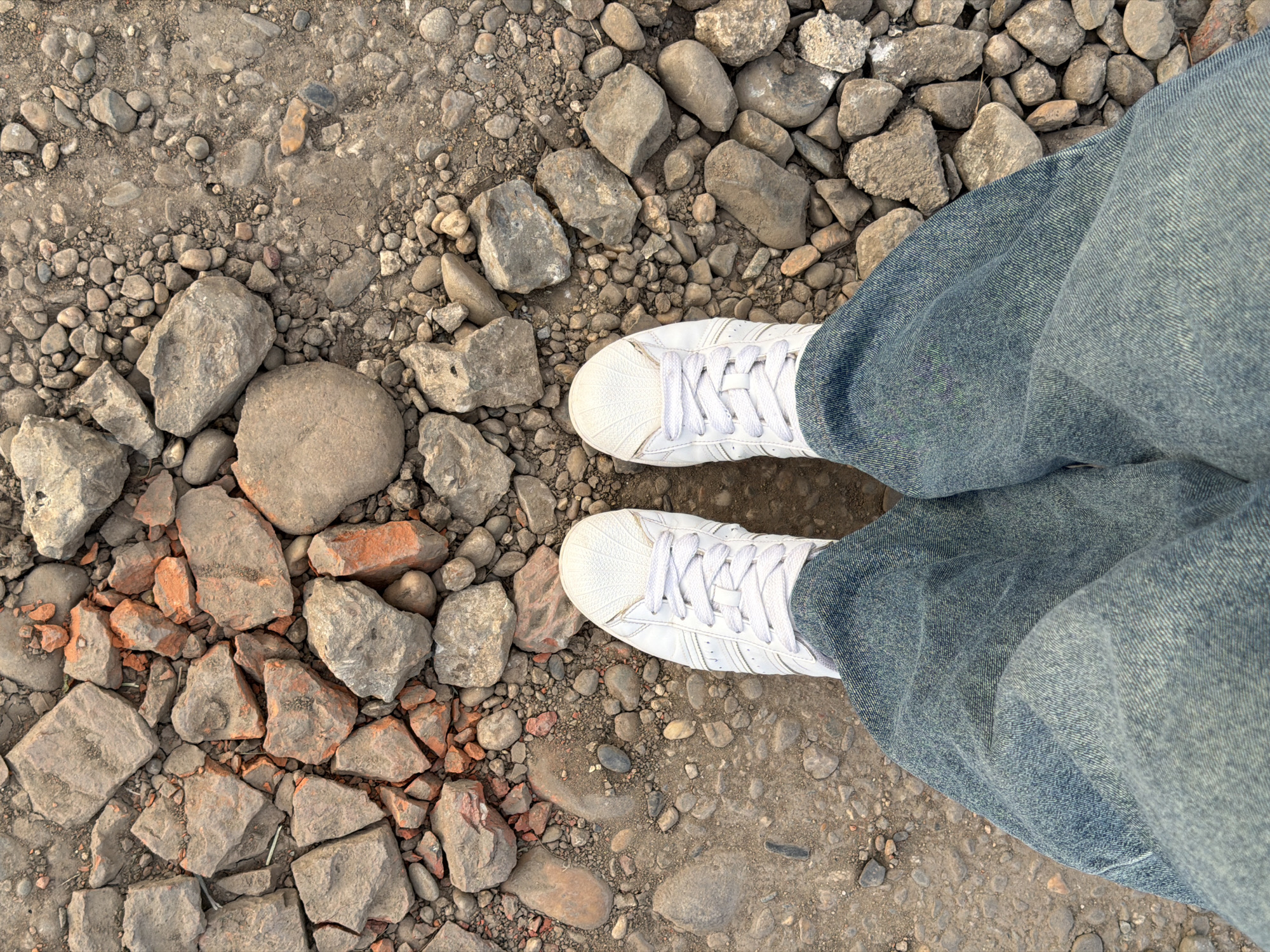 The image shows a downward view of a person standing on an outdoor ground surface composed of gravel, dirt, and broken rock fragments. The subject is wearing wide-leg, light-wash denim jeans and white sneakers with white laces. The location is inferred to be Rossiyskiy, Russia, based on the prompt, though there are no distinctive location markers visible within the image. No actions or interactions are depicted. The environment appears to be an unpaved area.