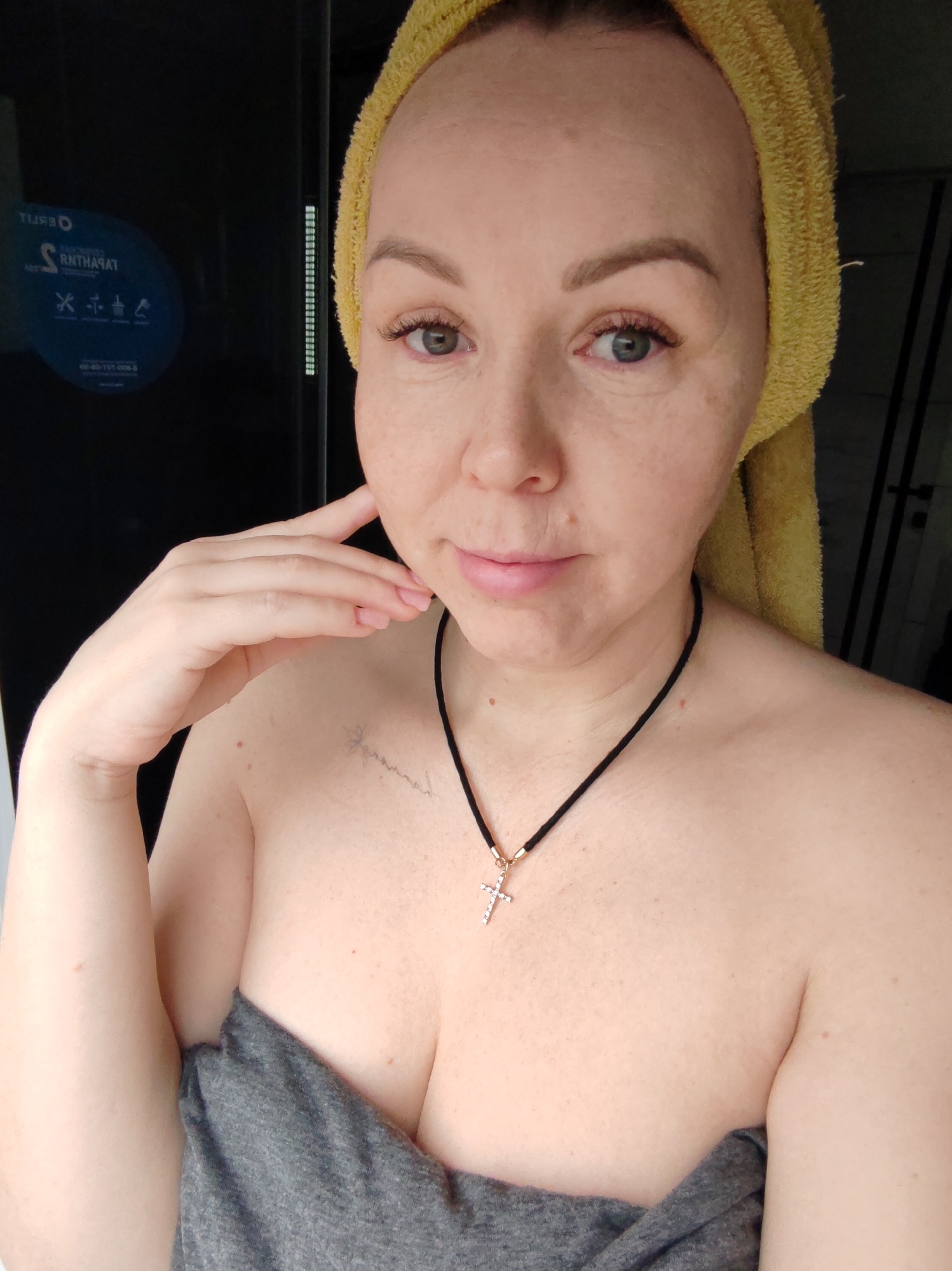A woman with fair skin is centered in the frame, likely indoors. A yellow towel is wrapped around her head. She is wearing a black necklace with a cross pendant. Her left hand is raised to her face, the fingers resting on her cheek. A gray garment appears to be draped around her torso. A tattoo is faintly visible on her chest. Behind her is a dark, possibly glass, partition with a circular sticker on the upper left. The background includes a hint of a tiled wall. The city is not discernible from the image.