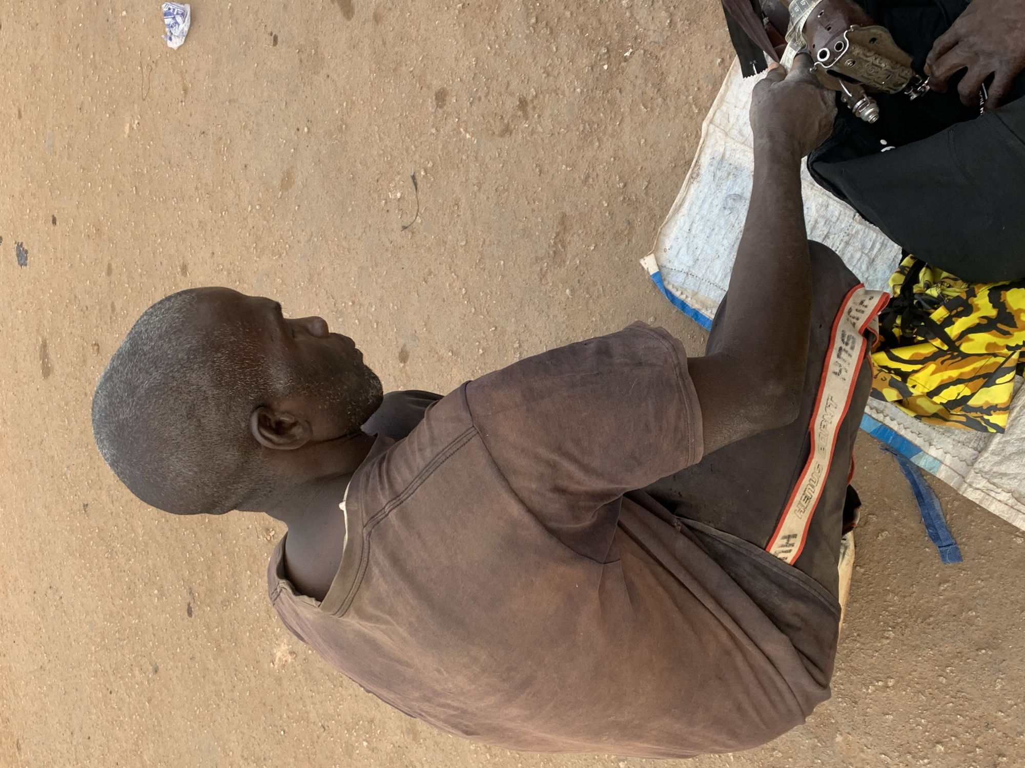 The scene depicts a man, viewed from above, seated on a patch of ground in an unidentifiable location within the city of Jos, Nigeria. The ground is light brown. He is wearing a faded brown t-shirt and dark shorts featuring a red and white stripe down the side. He is operating a small, hand-operated sewing machine on a piece of black cloth, which is resting on a white cloth spread out on the ground. The setting appears to be an informal workspace, and the man appears to be engaged in a sewing task. A swatch of yellow and black patterned fabric is also visible on the white cloth.
