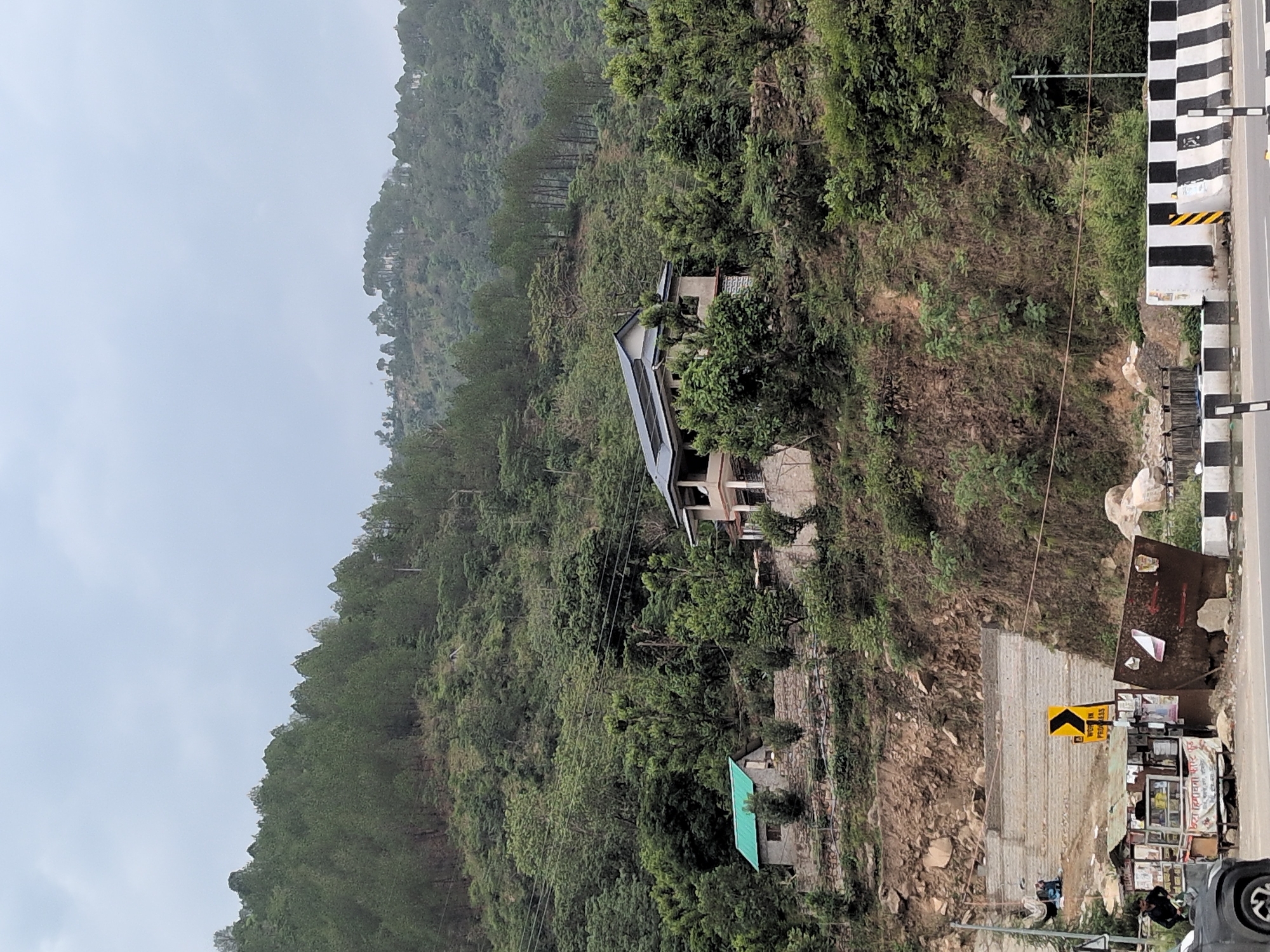 The image depicts a hillside scene in a mountainous region, likely in Sambal, India. A dense forest of pine trees covers the upper slopes of the hill. Below the forest, there are two houses situated on the hillside. The larger house, with a light-colored facade and a dark-tiled roof, is built on a terraced section of the slope. To the left and slightly lower down, a smaller house with a green roof is visible.
The foreground shows a roadside area. A road with black and white striped barriers runs horizontally across the bottom of the image. Adjacent to the road, there is a small roadside stall or shop with various items displayed. Several people are visible near the stall, including one person standing to the left and others in the background. A yellow road sign with a black arrow indicating a turn is also present. The ground in front of the stall appears to be a construction or work zone, with some debris and materials scattered around. A grey car is partially visible on the far left. The overall environment suggests a rural or semi-rural setting with evidence of development and daily life along a roadway. The sky is overcast, indicating a cloudy or possibly misty day.