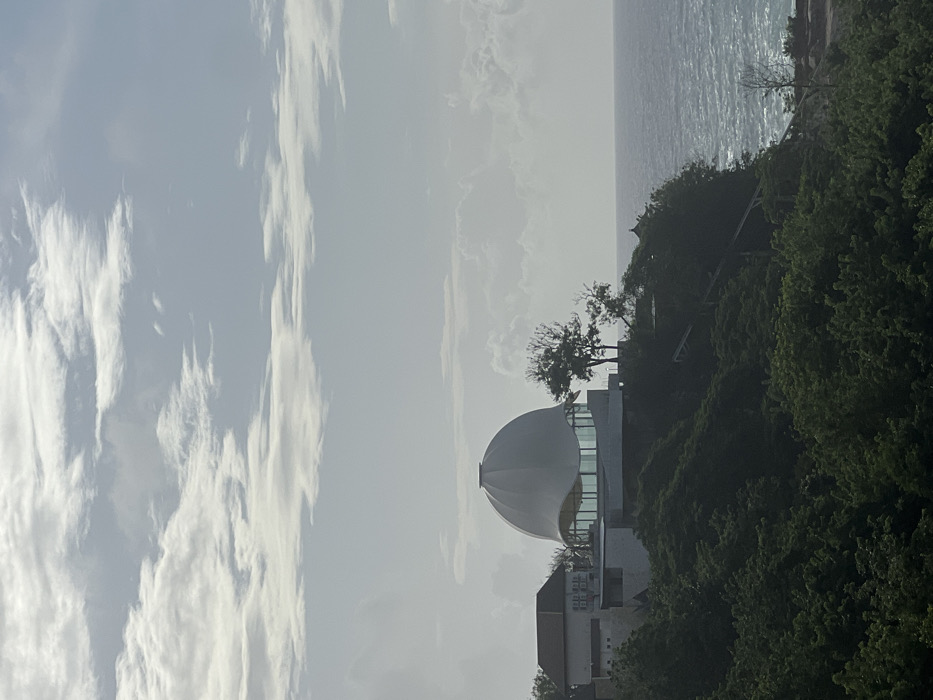 The image displays a panoramic view of the coastline in Uluwatu, Indonesia, under a partly cloudy sky. The primary subject is a distinctive modern building complex situated atop a verdant cliff on the right side of the frame. The central structure features a large, smooth, white, ovoid dome that appears to be the roof of a circular building. Beneath the dome, a section of the building has extensive curved glass windows, reflecting the sky. To the immediate left of this domed structure, a portion of another building with a traditional tiled or gabled roof and light-colored walls is visible.
The buildings are surrounded by dense, dark green foliage covering the cliff face. A few sparse, deciduous-looking trees are visible near the domed building and further down the cliff. On the right side of the image, the blue-gray ocean stretches to the horizon, with visible whitecaps indicating active waves near the shore. A faint, low-lying structure or rock formation is discernible at the water's edge on the far right. The sky occupies approximately the upper third of the image, displaying a mix of bright white cumulus and stratiform clouds against a lighter blue-gray haze. No individuals or specific actions are visible in the scene.