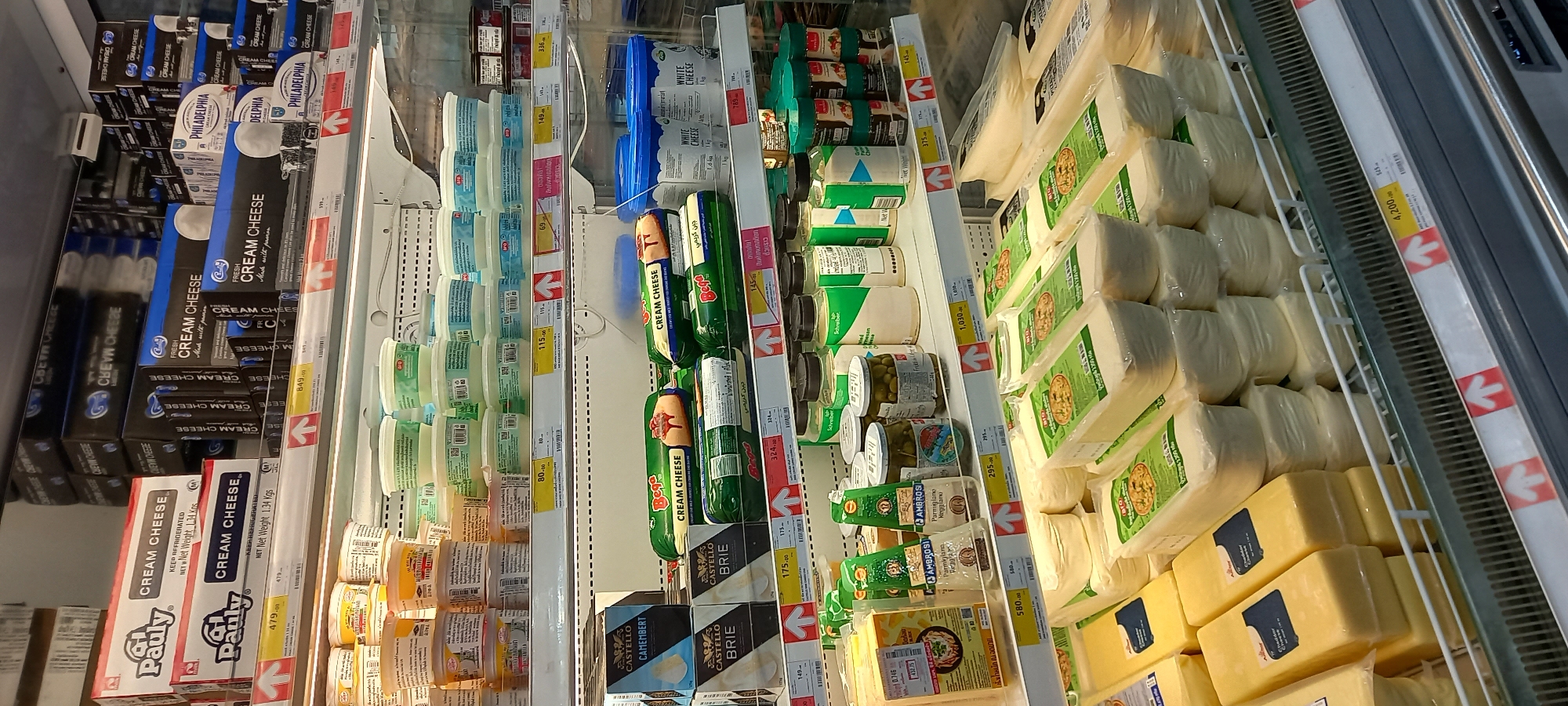 The media file captures a vertical view of a refrigerated display case in a supermarket or grocery store in Moo 1, Thailand. The shelves are filled with various dairy products, predominantly different types of cheese.
The top left section displays multiple boxes of cream cheese, including Philadelphia brand and a brand named Pauly, with some boxes noting "Net Weight 1.34 Kgs." Below these are more cream cheese boxes. To the right, numerous plastic tubs of various dairy spreads or yogurts are stacked.
The central part of the cooler features more cream cheese products, including tubes of Bega Cream Cheese and other brands, alongside small rectangular blocks of white cheese in blue tubs. Further down, there are small jars of spices or seasonings, and various types of pickled items like olives and gherkins in glass jars.
The right side of the display showcases large blocks and smaller wrapped portions of hard cheeses, possibly mozzarella or cheddar, many with green-labeled packaging. At the bottom, several rectangular blocks of yellow cheese are visible, some wrapped in clear plastic. Brands such as Castello (Brie, Camembert) and Ambrosia (Parmigiano) are also present.
Numerous price tags are affixed to the shelves, displaying prices in Thai Baht, ranging from 43.75 to 4,200. Some price tags feature red arrows, often indicating sales or promotions. The text "CREAM CHEESE," "FRESH CREAM CHEESE Made with passion," "WHITE CHEESE," "BRIE," "CAMEMBERT," and "Parmigiano" are clearly visible on product packaging. The lighting is typical for an indoor retail environment.