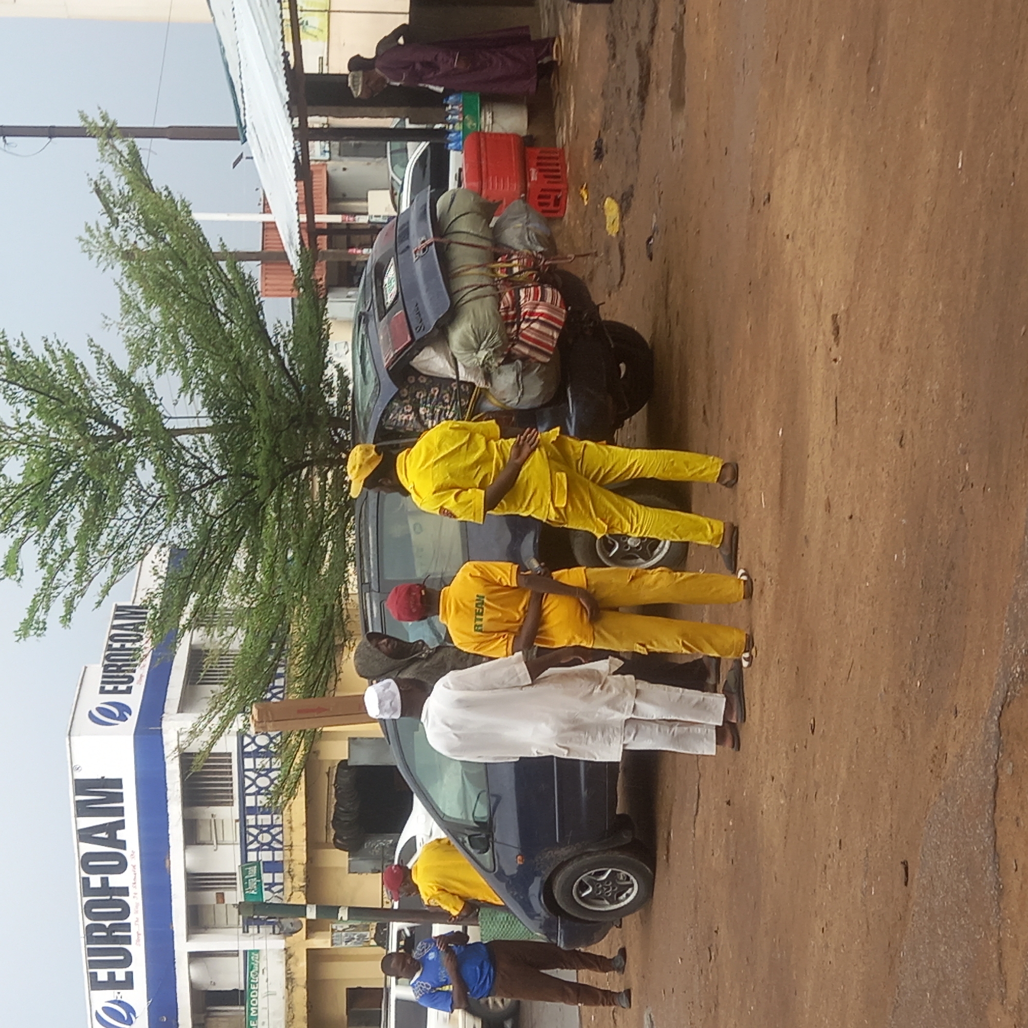 A vertical, medium shot depicts a roadside scene in Dadi Riba, Nigeria. The ground is brown dirt. A blue Eurofoam building stands to the left, partially obscured by a tree. A dark blue car, heavily loaded with bags and tires strapped to the roof, is parked next to a group of five men. Two men wear yellow outfits with red baseball caps, one with “RTEAM” printed on the shirt. Two other men wear matching yellow outfits. The last man, dressed in a white robe and cap, stands beside the car. Another woman is standing under the shade of the building. A man in a blue shirt and brown pants is also standing by the car.