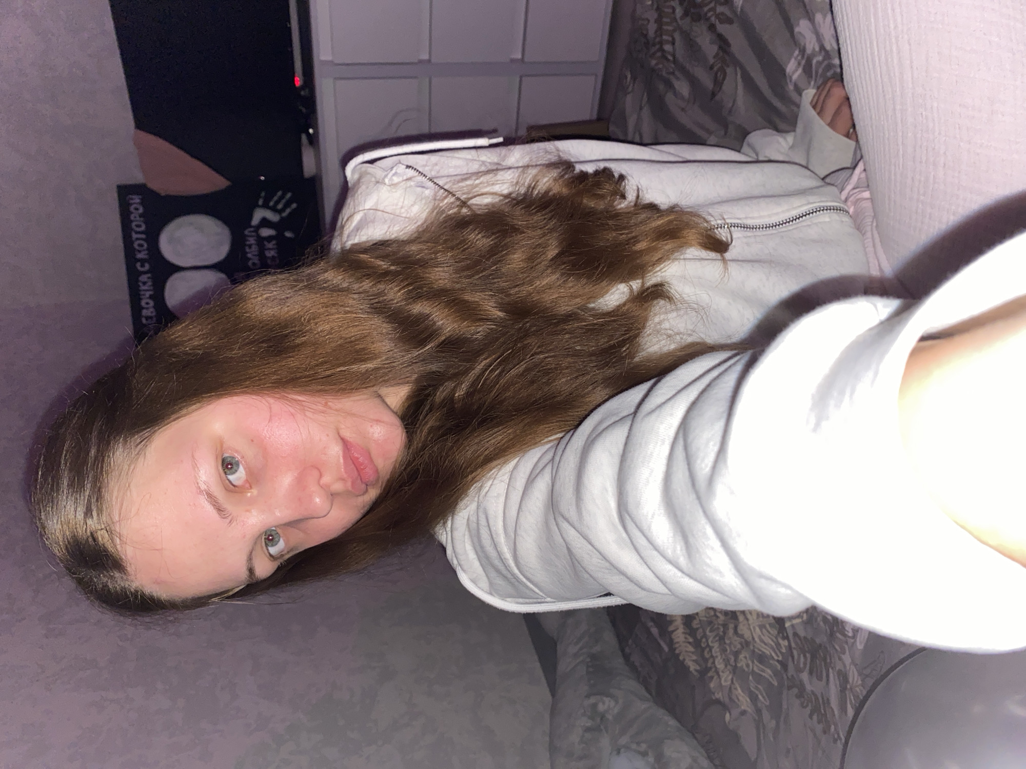 The image shows a young woman with long brown hair and light blue eyes, wearing a white zip-up hoodie and light pink pants. She is sitting on a bed with patterned bedding and a light pink blanket. To her right, a black canvas with images of the moon and text is visible, and a portion of a dark television screen is above white drawer units. The lighting in the room appears to be dim, with a purple hue. The overall scene suggests an indoor setting, possibly a bedroom, in a residential environment.