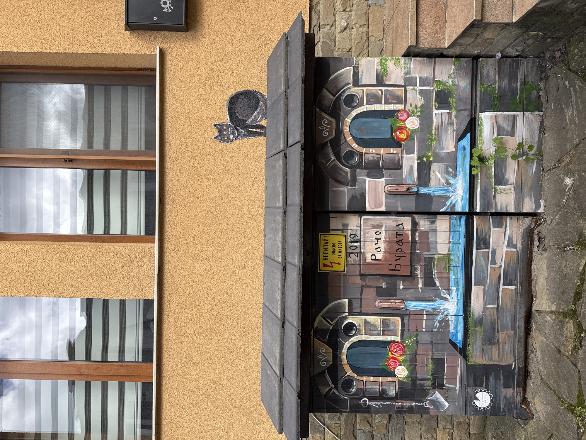 This image depicts a street scene in Gabrovo, Bulgaria. An ochre-colored building with two windows, each covered with slatted blinds, is visible on the left. A stylized black cat painting is present on the building's exterior. Adjacent to the building, a metal electrical box is painted with an elaborate mural depicting a water fountain motif, complete with floral elements, faux stone blocks, and a painted stream of water that extends across the two doors of the box. A sign written in Bulgarian and containing an electrical hazard symbol is affixed to the box. The words "Рачо Бурата" are painted above the sign. The box sits on a paved surface, which transitions to a stone staircase.