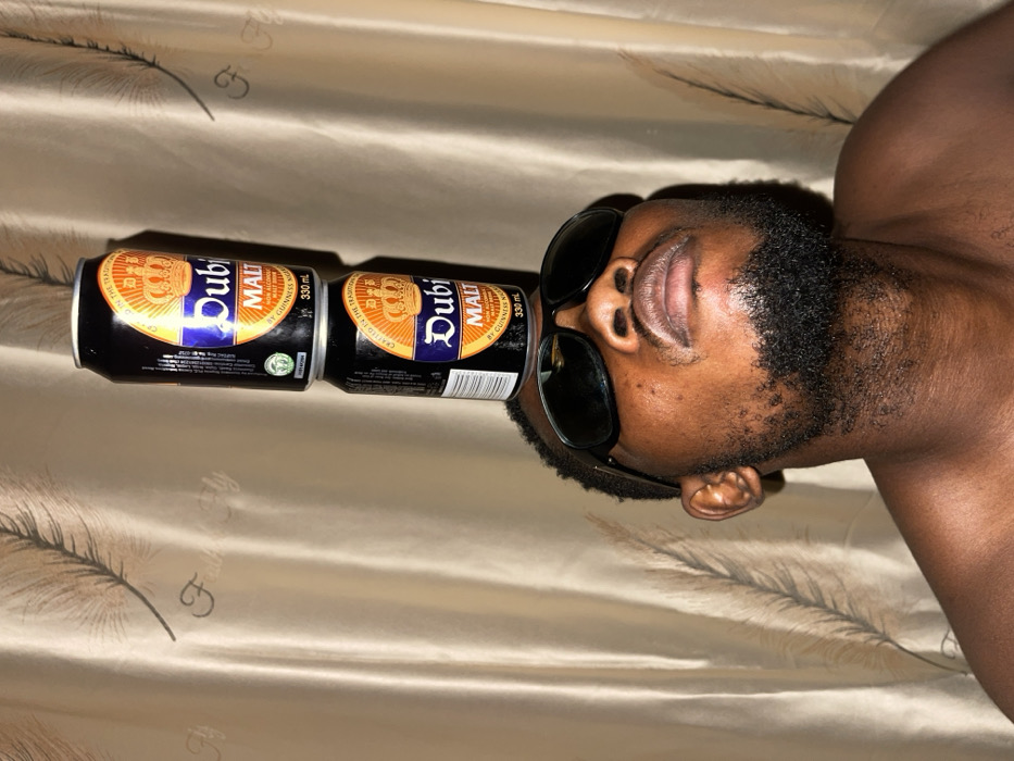 A man wearing sunglasses balances two Dubi Malt cans on his face. The cans, lined up in a row, rest on his eyes, supported by the sunglasses. The cans feature a gold crown design and product information in black and white text. The man has dark skin, a beard, and exposed shoulders. The background consists of a shiny, textured beige fabric with a feathery pattern. The image is oriented horizontally.