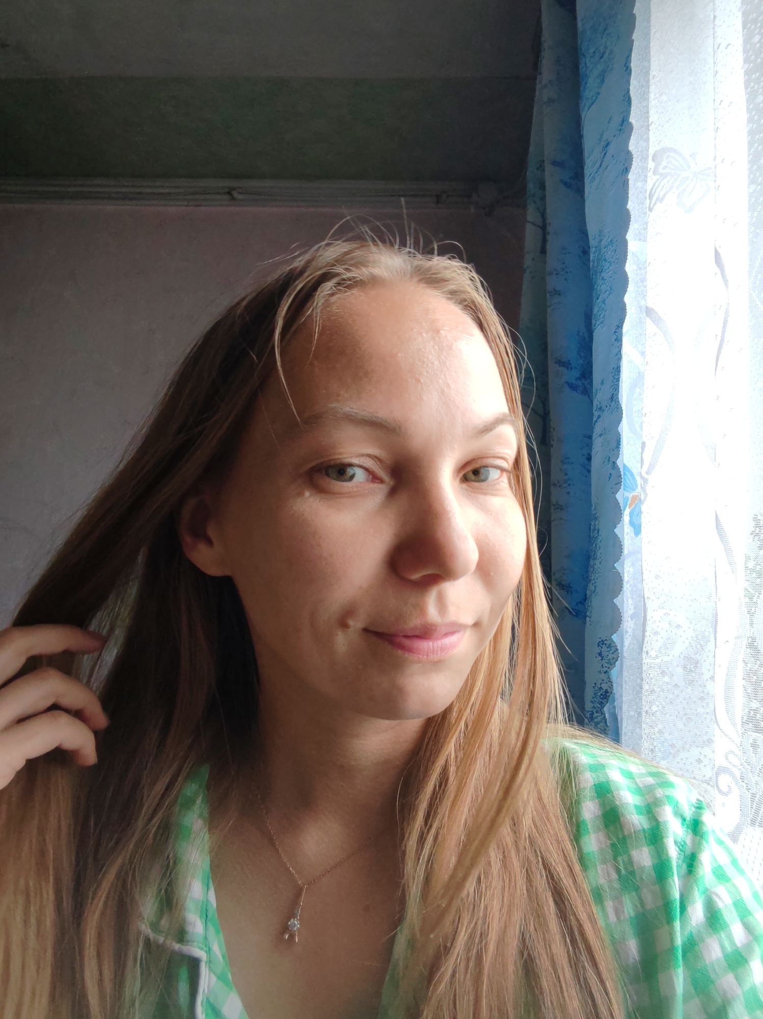 A young woman with long, blonde hair is shown from the chest up, posing for a selfie in front of a window with blue and white curtains. She is wearing a green and white checkered shirt and a delicate necklace with a pendant. Her right hand is brushing her hair aside. The background appears to be an indoor setting, possibly a room in a residential building in the city of Chukhlem, Russia, suggested by the style of the curtains and the general appearance of the room. The lighting indicates it is daytime, with natural light coming from the window.