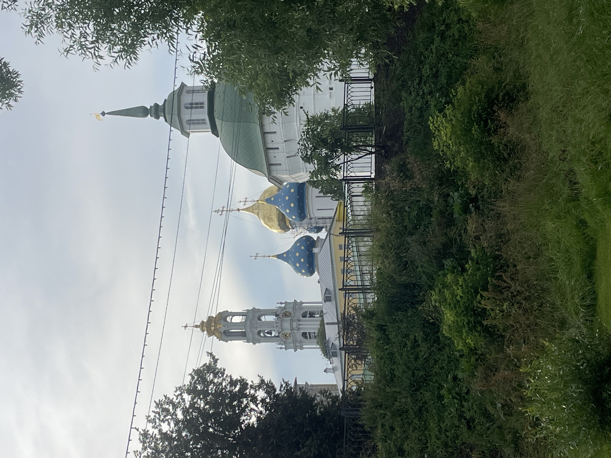 The image shows a view of the Trinity Lavra of St. Sergius in Sergiyev Posad, Russia. In the foreground, there is lush green vegetation, including trees and bushes, obscuring a black metal fence. Behind the fence, several church structures are visible. To the left, a tall, white bell tower with a golden spire stands prominently, featuring multiple arched openings and clock faces. Adjacent to the bell tower are church domes. One dome is bright blue with gold stars, topped with a cross, and another is golden. Further to the right, a large white building with a green domed roof is visible. The sky is overcast with some light clouds. The overall scene depicts a religious complex partially framed by nature.