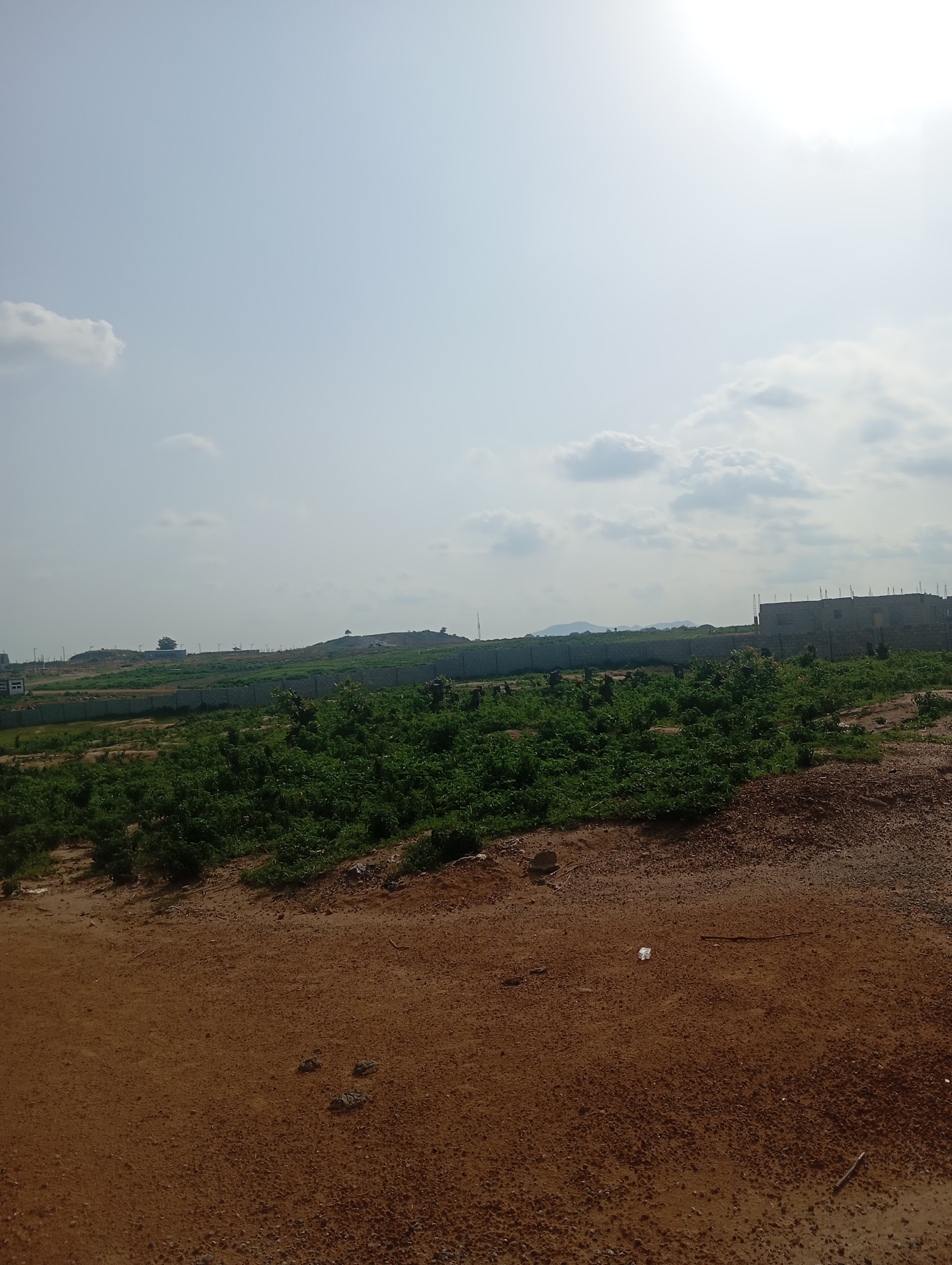 The image depicts an outdoor scene in Karsana 2, Nigeria, characterized by a construction site and surrounding undeveloped land. The foreground consists of reddish-brown, dry earth with scattered small stones and pebbles. A white plastic bag and a thin stick are visible on the ground in the mid-foreground.
In the mid-ground, a densely vegetated area with green shrubs and plants covers the landscape. This vegetation extends towards a low, stone-walled perimeter that encloses the area. Beyond the wall, the terrain rises into gently sloping hills.
The background features a large, multi-story building under construction, made of grey concrete blocks, with exposed rebar at the top. Further in the distance, other buildings and structures are visible, along with more hills and mountains on the horizon. The sky is overcast with scattered clouds, and the sun is faintly visible, creating a bright, washed-out effect in the upper right portion of the image. The overall impression is one of ongoing development in a semi-rural or exurban setting. There are no people or animals visible in the frame.