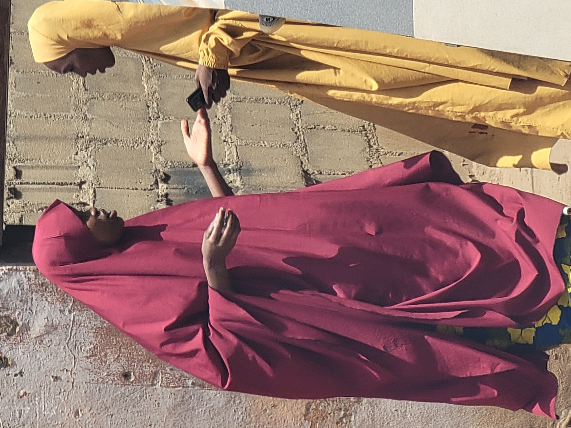 The photograph, taken in Maiburiji, Nigeria, depicts an interaction between two individuals in bright daylight, casting strong shadows.
The upper figure, likely a woman, wears a vibrant yellow head covering and a matching loose, long-sleeved garment. Her face is visible in profile, looking towards the other person. She holds a dark, rectangular smartphone in her right hand, positioned as if to hand it over or show it.
The lower figure, also likely a woman, is adorned in a flowing, voluminous maroon or magenta head covering and matching attire. Her face is turned upwards, gazing at the first person. Her right hand is raised and slightly open, seemingly in a gesture of reaching, receiving, or sharing in the interaction related to the smartphone. Her left hand is also visible, resting folded.
The background features a textured, light-colored brick or concrete wall behind the yellow-clad person. Below that, a lighter, rough-textured surface, possibly ground or another wall, is visible. Hints of a colorful blue and yellow patterned fabric peek out from beneath the maroon garment on the right side. On the yellow garment, near the shoulder, a small, faint white logo or tag, possibly a stylized 'M', is visible. The strong sunlight and clear visibility indicate a sunny day.