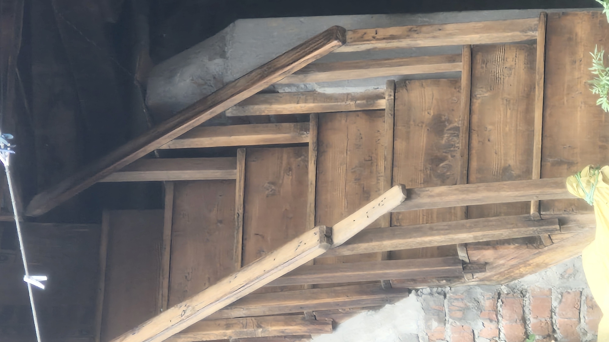The image depicts a rustic interior scene, likely an attic or a storage area, characterized by exposed wooden beams and planks. A prominent diagonal wooden beam cuts across the frame from the lower left to the upper right, appearing to be a structural element or perhaps a railing. Beneath this beam, more horizontal wooden planks form a sort of layered or tiered structure. The wood is weathered, showing grain patterns and some signs of wear, suggesting age and a non-modern construction.
In the background, a dark, shadowy area is visible, possibly a void or an unfinished section of the building. To the right, a patch of green foliage from a plant peeks into the frame, and a portion of a yellow fabric, possibly a cloth or a garment, is draped and secured with a green string. The lower part of the image reveals a rough, unfinished wall made of brick and mortar, further reinforcing the rustic and possibly utilitarian nature of the space. The lighting suggests an indoor environment, possibly with natural light filtering in from an unseen source, creating areas of light and shadow. No people are visible in the frame. The overall impression is one of a simple, aged structure, potentially in a rural or traditional setting. Given the location context of Vashisht, India, this could be part of a traditional Indian dwelling or outbuilding. There is no visible text in the image.