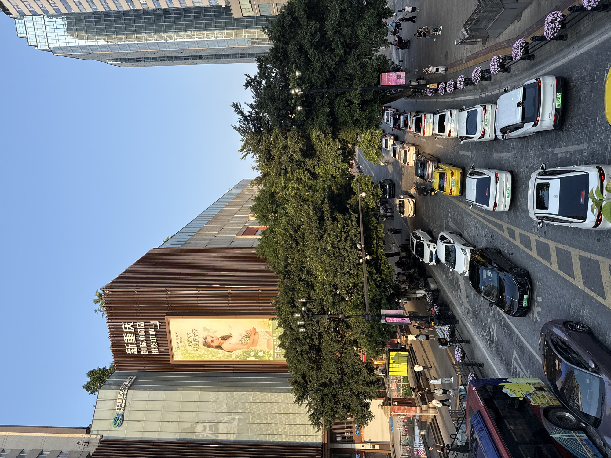 This image captures a bustling street scene in an urban area, likely Yuzhong District, China, as seen from an elevated perspective. The setting is a multi-lane road flanked by tall, modern buildings and lush green trees.
The primary activity is vehicular traffic, with numerous cars forming a queue or moving slowly along the street. Many of these vehicles, mostly white sedans and SUVs, have distinctive green license plates (e.g., AA BF3172, AA D74669, AA D3981, AA A00311), indicating they are New Energy Vehicles (NEVs). Other vehicles include a yellow car, a black car, a purple car, and a red and blue double-decker bus adorned with Minions graphics. People are visible on the sidewalks, walking, standing, or waiting, adding to the urban vibrancy.
Notable details include a prominent building with a brown slatted facade, featuring a large billboard displaying a woman in lingerie with the brand "DEANFUN" and Chinese text that translates to "Comfortable Underwear, it's Deanfun/Butterfly Comfort." Below the billboard, text reads "新重庆 国际小商品 批发中心" (New Chongqing International Small Commodities Wholesale Center). Adjacent to this, another building with a glass facade shows "中国民生银行" (China Minsheng Bank) and "CHINA MINSI". The street is lined with lampposts decorated with hanging baskets of purple flowers. The sky is clear and blue, suggesting a daytime scene, possibly late afternoon given the soft lighting. The overall impression is one of a busy, modern city environment.