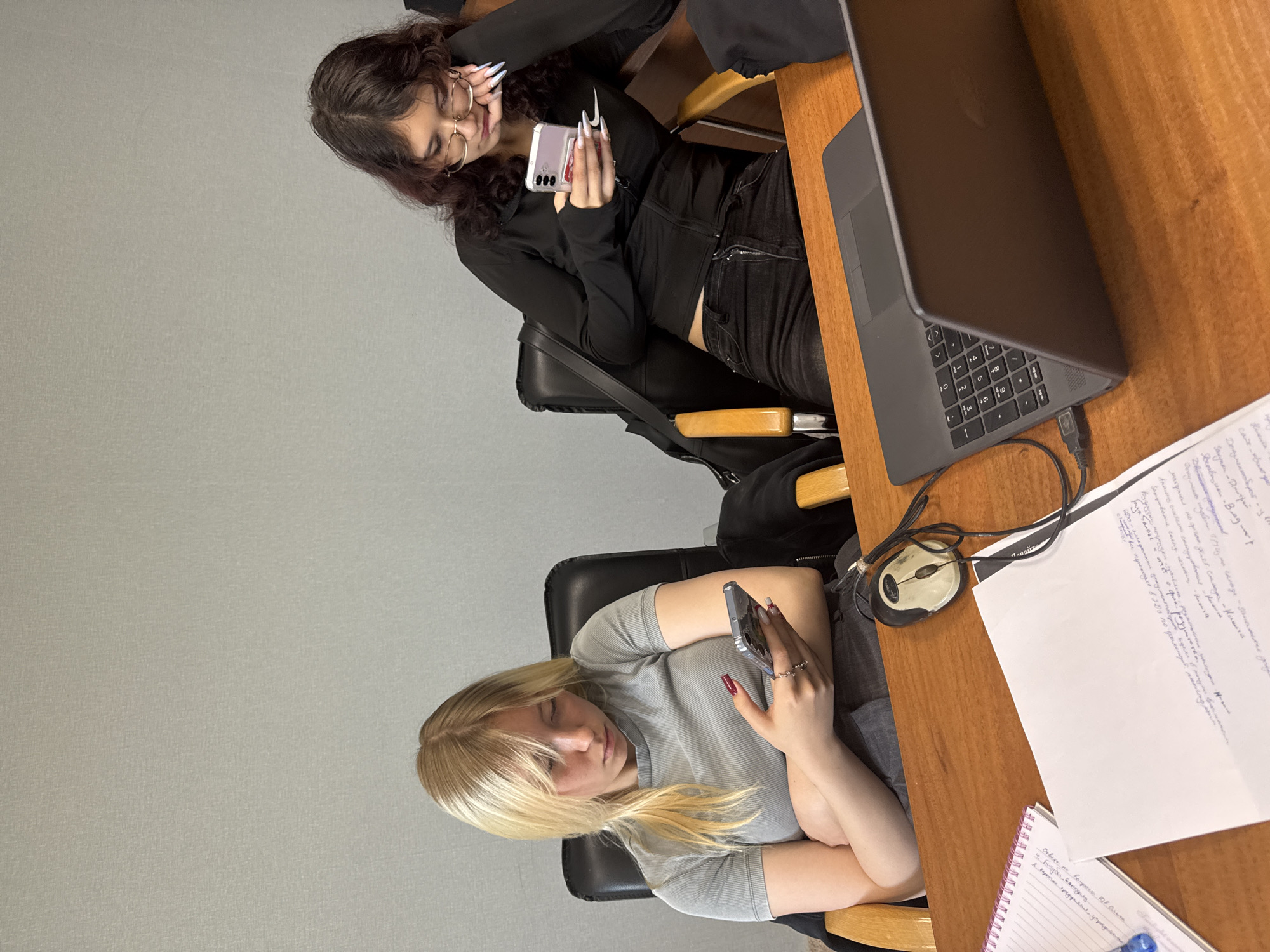 The image depicts two young women sitting at a wooden desk in what appears to be an office or study environment. The woman on the left has blonde hair and is wearing a grey t-shirt. She is holding a smartphone in her right hand and looking down at it. Her arms are crossed. The woman on the right has dark, wavy hair and is wearing glasses and a black zip-up jacket. She is also looking at her smartphone, which she holds in her right hand. Her left hand is resting on her chin, and her elbow is propped on the desk.
On the desk, there is a laptop computer, a computer mouse, a notebook with writing on it, and loose papers with handwritten text. The background is a plain, textured grey wall. The overall lighting suggests an indoor setting. The presence of office furniture and equipment indicates a workspace. The image does not contain any specific visual cues that would allow for definitive identification of the location as being in the city of Perm, Russia.