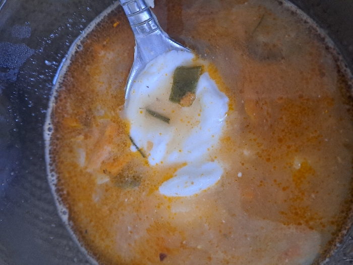 The image displays a close-up, top-down view of a reddish-orange liquid dish within a dark-rimmed container, located in the city of Bel'kovo, Russia. The surface of the liquid exhibits numerous small, orange fat droplets and a distinct frothy, dark-orange ring along the inner edge of the container. A metallic spoon is partially submerged in the liquid, holding a white, viscous substance, consistent with dairy, along with several dark green fragments and a smaller, light-orange food particle. Additional light-colored and dark-green food particles are observed suspended within the main body of the liquid. The spoon's presence indicates an action of serving or consumption.