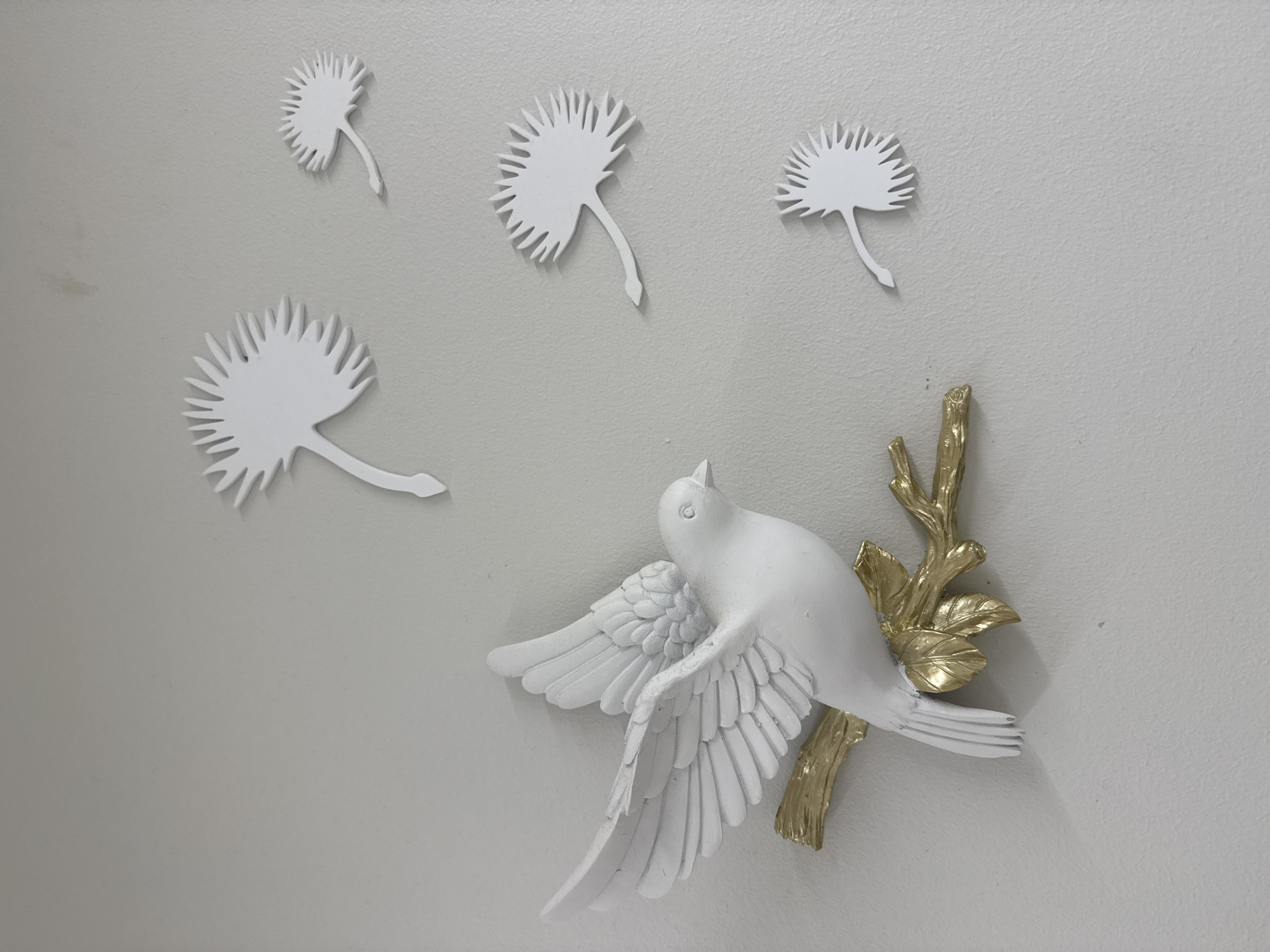 A close-up, angled shot depicts white, stylized decorations on a light gray wall. Four white, flower-like shapes, reminiscent of dandelions or palm fronds, are affixed to the wall above and to the left. Below and to the right, a white bird with outstretched wings appears to perch on a golden branch with leaves. The bird and branch are also attached to the wall. No people are visible. The location is not discernible from the image, and there is no indication it is in Riga, Latvia.