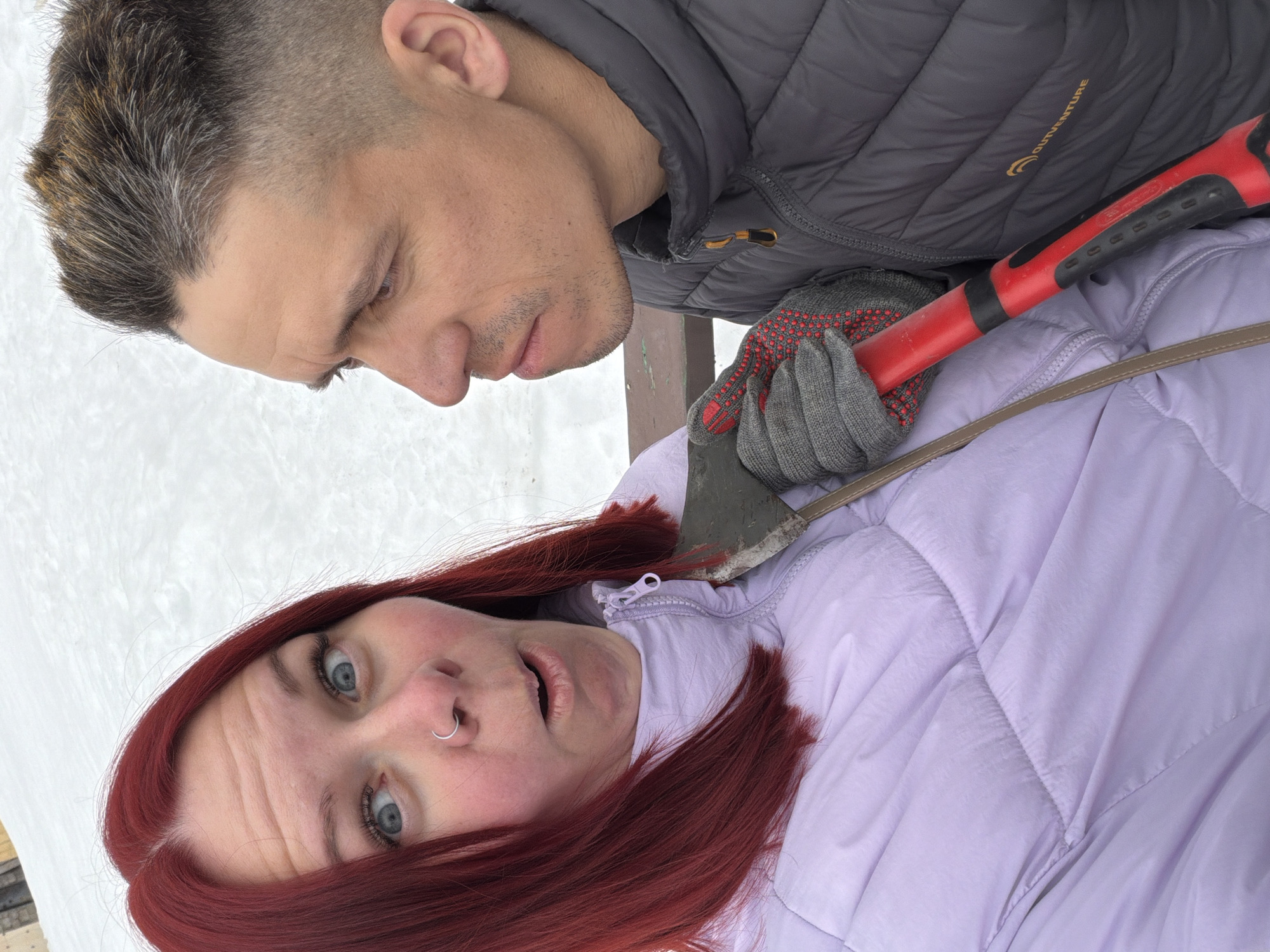 The image captures two individuals, one female and one male, in a close-up outdoor setting during winter. The ground in the background is covered in snow, and a blurred dark brown wooden object, possibly a bench or railing, is partially visible. The specific location within the city of Chaikovsky, Russia, is not discernible from the image.
The female, positioned on the left, has red hair, wide-open light blue eyes, and an open mouth, depicting an exaggerated expression. She wears a light lavender quilted winter jacket, zipped up, and a silver hoop nose ring on her left nostril. A light brown strap, possibly from a bag, is visible over her jacket.
The male, positioned on the right and slightly behind the female, has short dark hair with lighter highlights, minimal facial stubble, and dark eyes directed towards the female. He wears a dark grey quilted winter jacket, visible with a yellow "OUTVENTURE" logo on the left chest, and grey work gloves with red textured dots on the palms.
The male is holding a small axe with a red handle and a grey metal head in his gloved right hand. The blade of the axe is positioned near the female's upper chest/collarbone area, pointing towards her. Both individuals appear to be looking directly at the camera, suggesting a staged interaction.