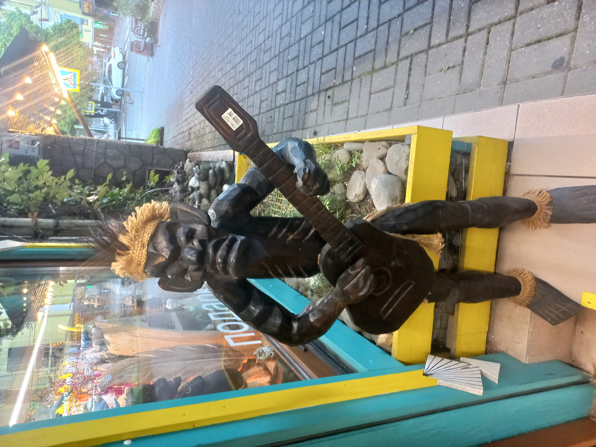 The image shows a wooden sculpture of a dark-skinned figure with a straw-like hair, positioned outside a store or shop in what appears to be a city setting. The figure is holding a wooden guitar, and the sculpture rests partially on a yellow-colored planter filled with rocks and some sparse greenery. The sculpture is in front of a glass window of the shop, which reflects the street scene, including a crosswalk sign, streetlights, and some buildings. The sidewalk is paved with interlocking concrete blocks. The location is suggested to be Sochi, Russia.