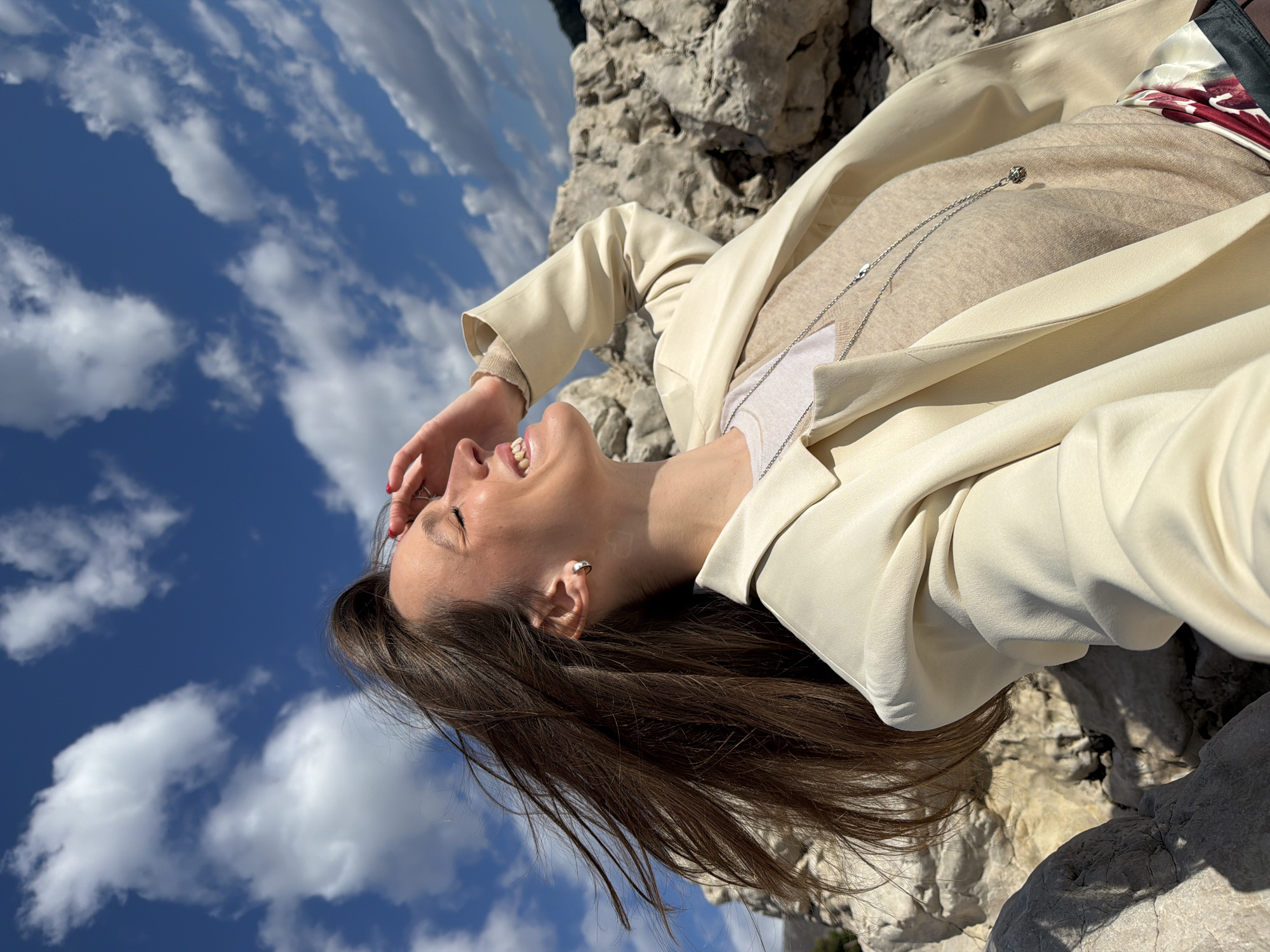 The image shows a young adult female positioned on light-colored rocky terrain in Sutomore, Montenegro, under a bright blue sky with scattered white cumulus clouds. The subject's head is tilted upwards and to her left, with her eyes closed or squinting, and her mouth open in a wide smile. Her long brown hair is visibly swept to her left, indicating wind. Her right hand, featuring red nail polish, is raised and positioned to shield her face from direct sunlight. She is wearing a cream-colored blazer over a beige long-sleeved top, with a white garment visible at the neckline. A silver-colored chain necklace with a small, spherical pendant hangs around her neck, and a silver hoop earring is visible in her left ear. The immediate environment consists of large, textured, off-white rock formations.