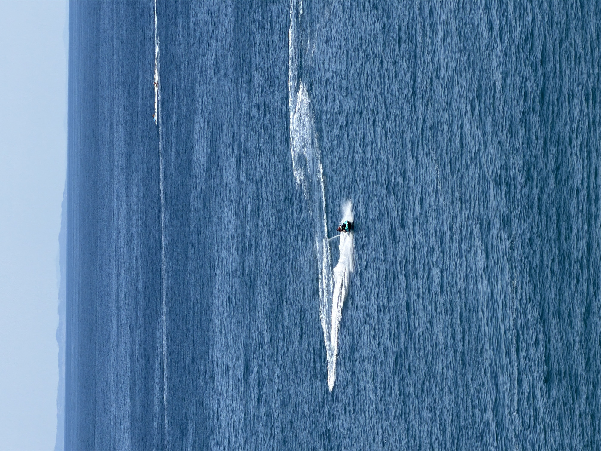An aerial view captures a vast expanse of the deep blue Adriatic Sea with a distant, hazy coastline of mountains and landforms. In the foreground, a person rides a jet ski, creating a prominent white wake that curves across the water. Further in the distance, another jet ski with two riders is visible, also leaving a smaller white trail. The sea is moderately choppy with a textured surface. The sky above is a pale, hazy blue, with a thin line of clouds near the horizon.