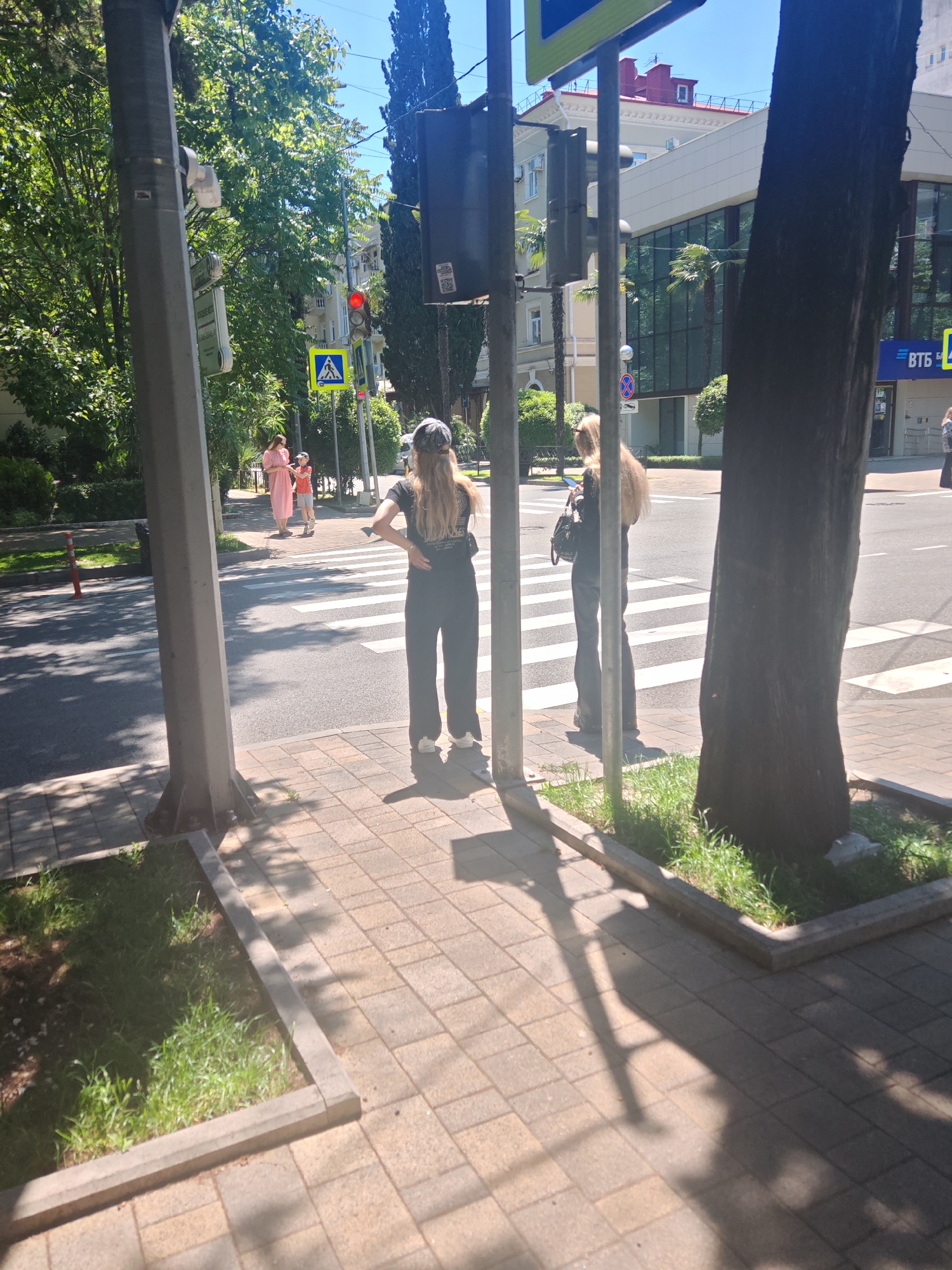 The image captures a sunny urban street scene, likely during mid-day or early afternoon due to the strong, defined shadows cast by trees and street furniture. The perspective is from a sidewalk, looking across a road towards buildings and trees.
In the foreground, a paved sidewalk with patterned pavers features a small, raised planter box with grass. On the left, a sturdy metal lamppost holds a security camera, various signs, and a traffic light. On the right, the thick, dark trunk of a large tree partially frames the scene.
Several people are visible, seemingly waiting to cross the street at a pedestrian crossing. Two young women with long, light-colored hair stand in the middle-ground on the sidewalk, facing away from the camera. One wears a black t-shirt, black wide-leg pants, white sneakers, and a backward baseball cap, with her hands on her hips. The other, dressed in black, holds a phone and a handbag. Further back on the left, near the same crosswalk, a woman in a long pink dress and a child in red shorts are also waiting.
The street features clearly marked zebra stripes for the pedestrian crossing. A red traffic light is visible, indicating that pedestrians should not cross. Below the red light, a small pedestrian signal with a white arrow is also present. A blue square sign with a white triangle and a black walking person symbol denotes the pedestrian crossing. Other road signs include a round blue sign with a red diagonal line ("No Stopping") and another round sign with a red border and a black truck icon ("No entry for goods vehicles").
In the background, the architecture varies from older buildings with ornate details and red roofs to more modern structures with large glass facades. Lush green trees line the street, including several palm trees, suggesting a warm climate.
Visible text includes "ВТБ" in Cyrillic script on the front of a modern building, which is a Russian bank. The red traffic light lens also appears to have a "STOP" graphic or pattern. Some text on a hanging sign to the left of the lamppost is illegible.