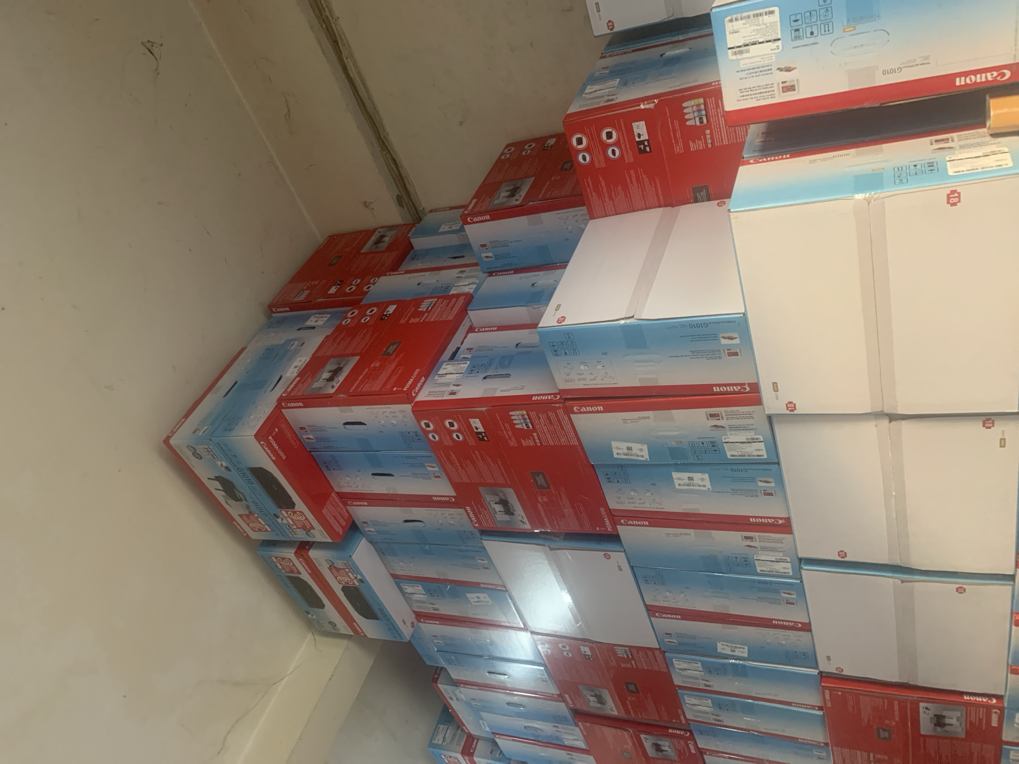 The image displays a large stack of Canon printer boxes, predominantly in red and blue packaging, filling a warehouse space. The boxes are stacked high against a light-colored wall, with a visible ceiling fixture or beam at the top. Some boxes feature prominent "Canon" branding and product model numbers like "G1010" and "PIXMA G1010." The overall impression is one of inventory storage. The context of Palembang, Indonesia, suggests this is a commercial storage facility or retail stockroom in that city. There are no people visible in the image, and the focus is solely on the stacked boxes.