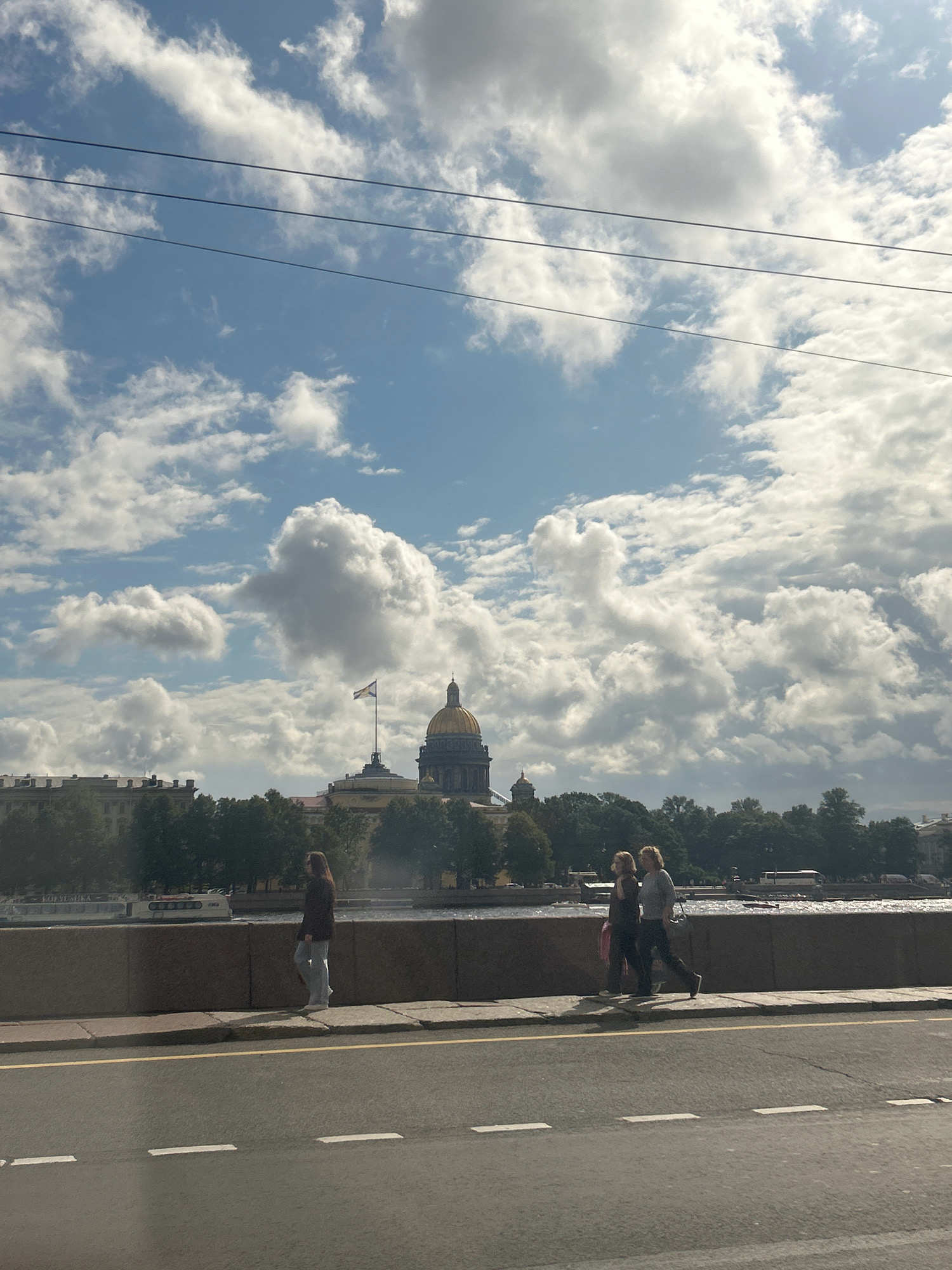 The image captures a view of St. Isaac's Cathedral in Saint Petersburg, Russia, under a cloudy sky. In the foreground, a road with white lane markings is visible. A concrete embankment lines the edge of the road, separating it from a sidewalk where three individuals are walking. To the left, a woman walks alone, facing away from the camera. Further to the right, two people, appearing to be a couple, walk side-by-side. Beyond the embankment and sidewalk, a body of water, likely the Neva River, is present, with a boat docked along its bank. Trees form a green line along the river, and St. Isaac's Cathedral, with its distinctive golden dome, is prominent in the background. A flag flies from a pole in front of the cathedral. The overall scene suggests a daytime urban setting with a mix of architectural landmarks and everyday life.