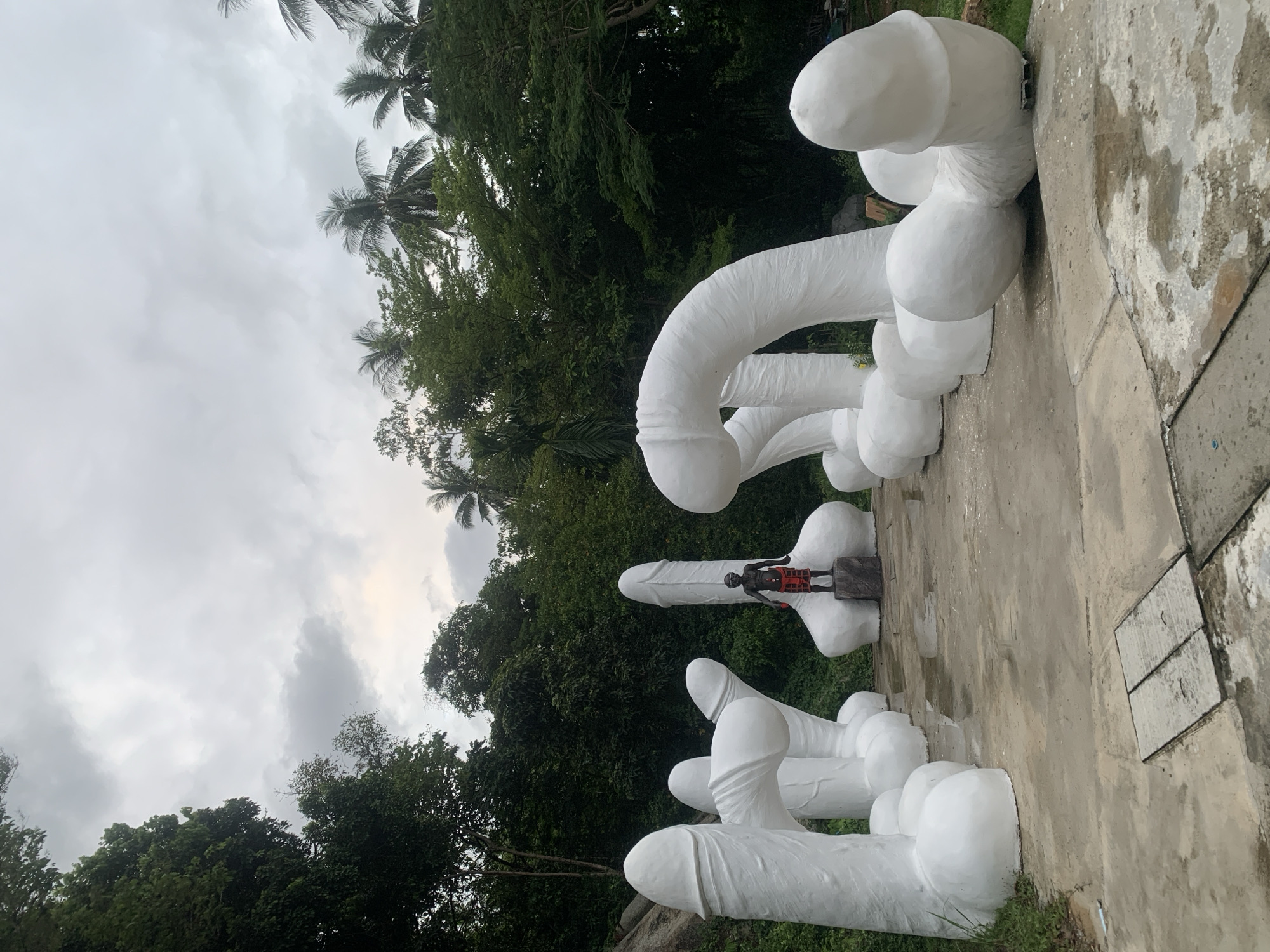 The image displays an outdoor scene featuring numerous large, white, phallic-shaped sculptures. These sculptures are of varying sizes and forms, arranged on a concrete platform, creating a prominent display in the foreground and midground. Some of the sculptures stand upright, while others are curved or clustered together.
Perched on top of one of the taller phallic sculptures in the midground is a small figurine. This figurine appears dark-skinned and is dressed in red shorts or a loincloth, standing erect with its arms slightly bent.
The background is filled with dense, green tropical foliage, including several tall palm trees whose fronds are visible against the sky. The sky is heavily overcast with grey clouds, indicating a cloudy or gloomy day. The concrete ground below the sculptures shows signs of moisture or staining. The overall setting, combined with the distinct nature of the sculptures, suggests a cultural or tourist attraction, likely a fertility shrine or a similar site, consistent with the provided location context of Baan Hua Thanon, Thailand. No visible text is present in the image.