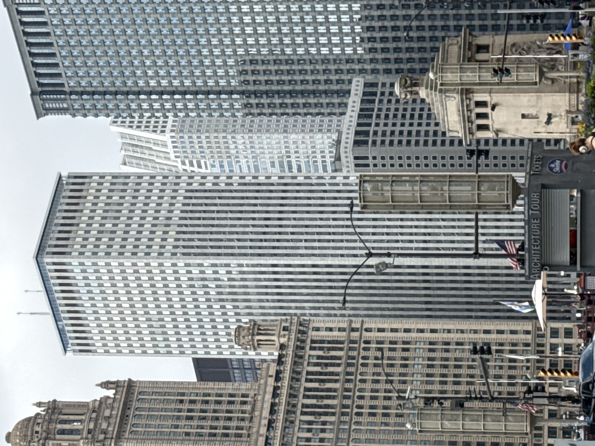 This media file captures a view of several tall buildings in an urban setting, likely Chicago. The architecture varies, with some buildings featuring a repetitive grid of windows and others exhibiting more ornate, classical designs, including domes and intricate detailing. The perspective is angled upwards, emphasizing the height of the structures. The sky appears overcast, suggesting a cloudy or hazy day.
Among the buildings, there are various objects visible. Traffic lights are present, indicating a street-level view or an intersection. Flags, including American flags, are displayed on some buildings. There are also signs, one of which reads "ARCHITECTURE TOUR" and another with "TICKETS" and a "Sears" logo, suggesting a tour bus or information kiosk. The presence of cars and what appears to be a tour bus hints at an active tourist area. Though not the primary focus, there are glimpses of people in the background, and a person's head with brown hair is visible in the lower right corner, possibly taking a photo or observing the scene.
The overall impression is of a bustling city environment, rich in architectural history and offering attractions for visitors. The time of day is not definitively clear due to the overcast sky, but it appears to be daytime.