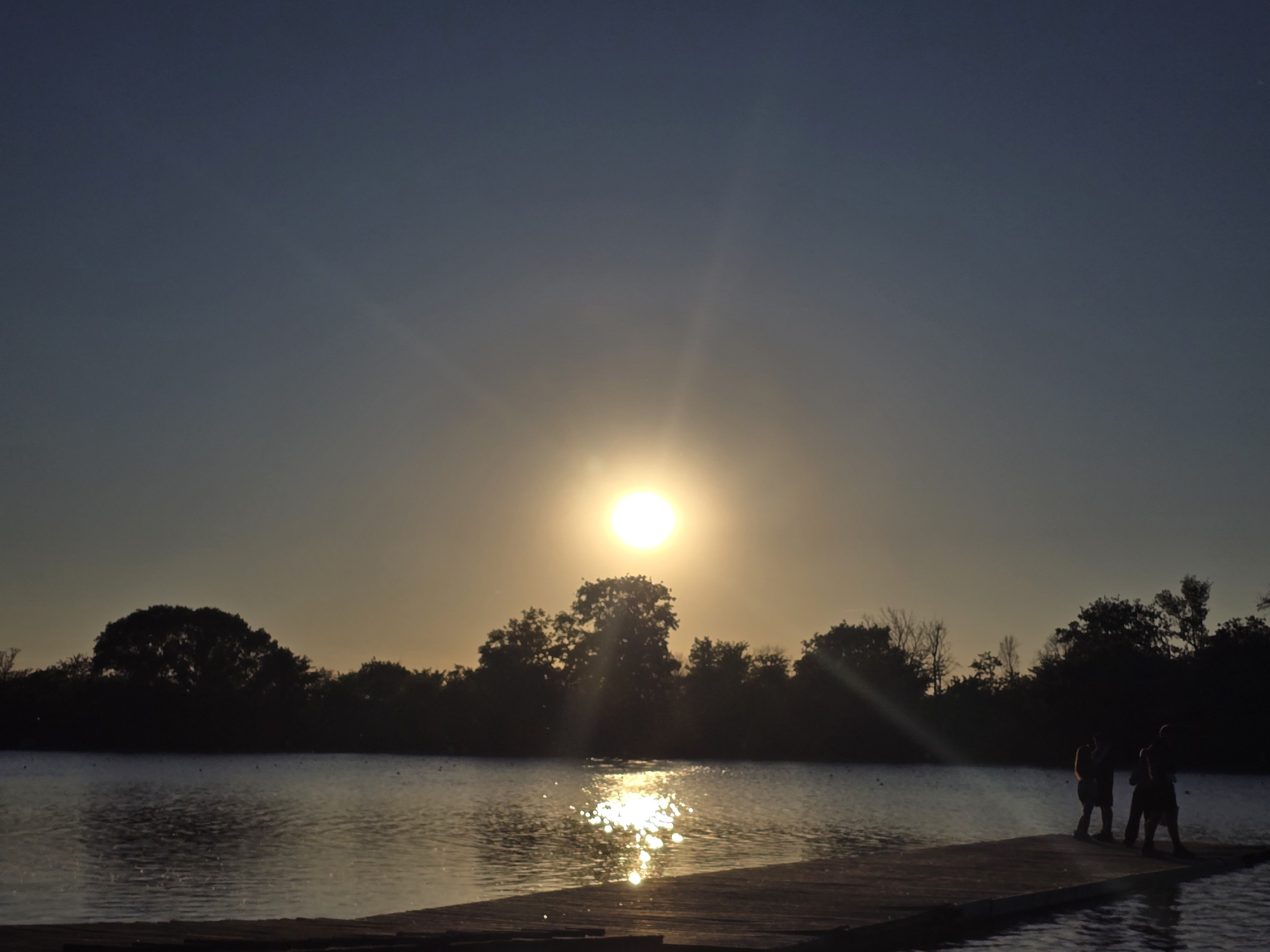 A lake scene at sunset in Unknown, Unknown. The sun is positioned high in the sky and is reflecting on the water's surface. A group of three people are standing on a dock. Trees line the shore of the lake. The sky is a gradient of blue and yellow hues.