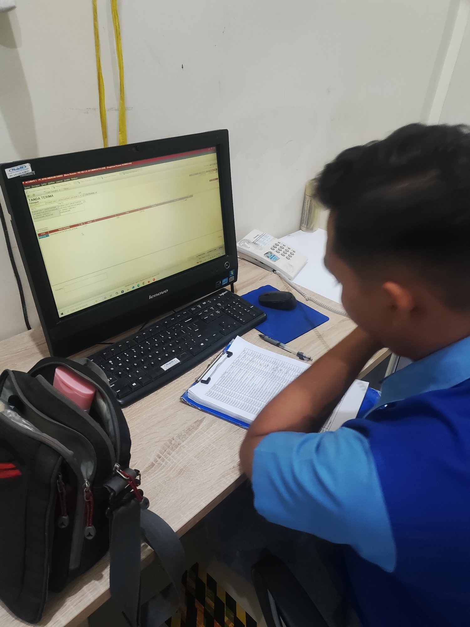 A young Indonesian man, wearing a blue collared shirt, sits at a wooden desk in what appears to be an office environment in Pepelegi, Indonesia. He is facing a computer monitor, which displays a spreadsheet or database with columns and rows of text, predominantly in red and white. A black keyboard and a black computer mouse on a blue mousepad are positioned in front of the monitor. To the right of the keyboard, a white corded telephone is visible. To the left of the keyboard, a black messenger bag with red accents is open, revealing a pink container inside. A clipboard with several documents is placed on the desk in front of the man, and he appears to be reviewing them while looking at the computer screen. The background consists of a plain white wall with two yellow cables running vertically along it.