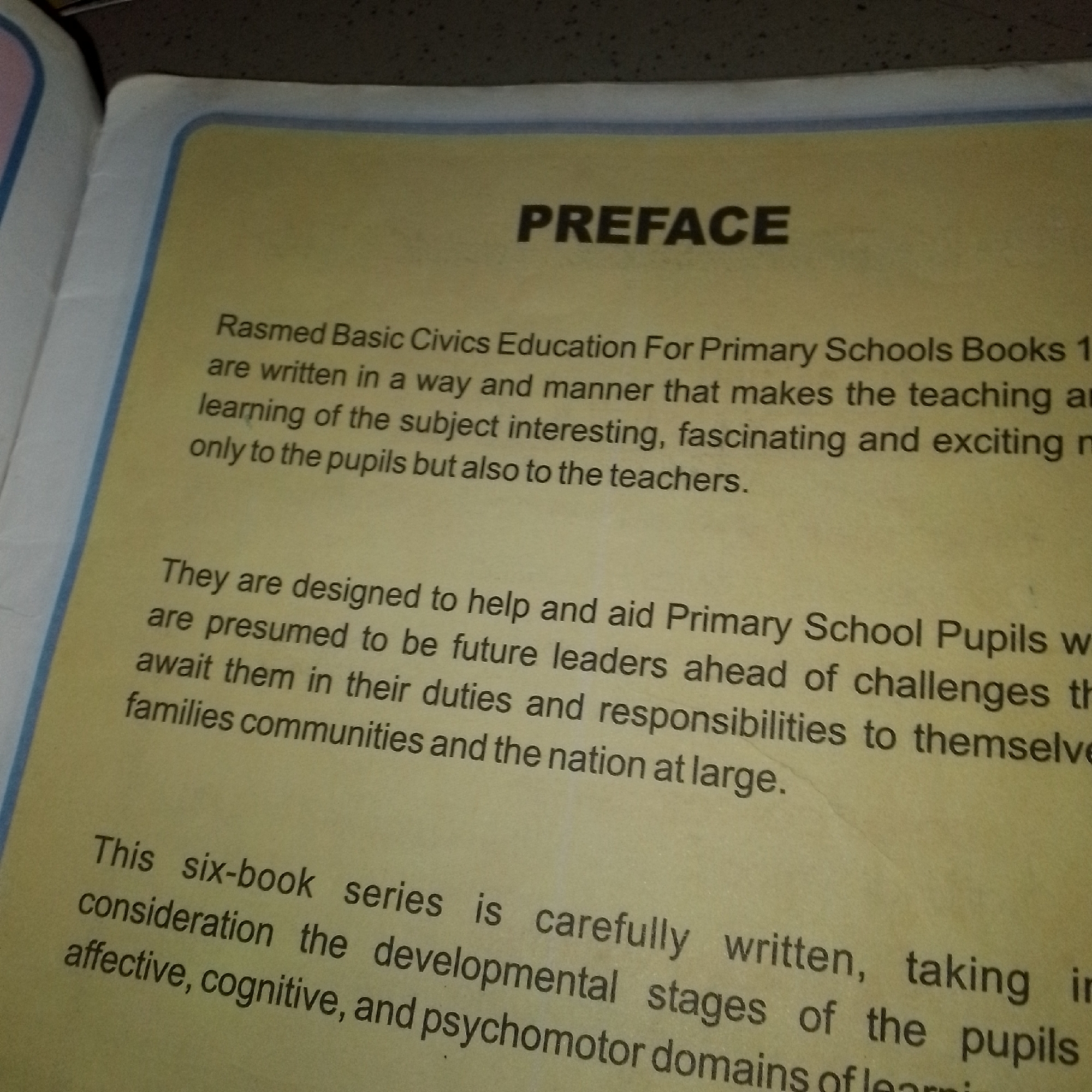 The image displays the preface of a book with the title "PREFACE" prominently centered at the top in bold, black capital letters. Below the title, the text is printed in black ink on a yellow-toned paper background, possibly aged. The text discusses the writing and purpose of a series of "Rasmed Basic Civics Education For Primary Schools Books." It highlights that the books are designed to make learning interesting and exciting for pupils and teachers, and to prepare future leaders by outlining their duties and responsibilities to themselves, families, communities, and the nation. The preface also mentions that the six-book series takes into consideration the affective, cognitive, and psychomotor domains of learning. The lighting suggests the image was taken indoors, with the book open on a surface. There are no people visible in the image. The geographical location of Jalingo, Nigeria, is not indicated in the image.