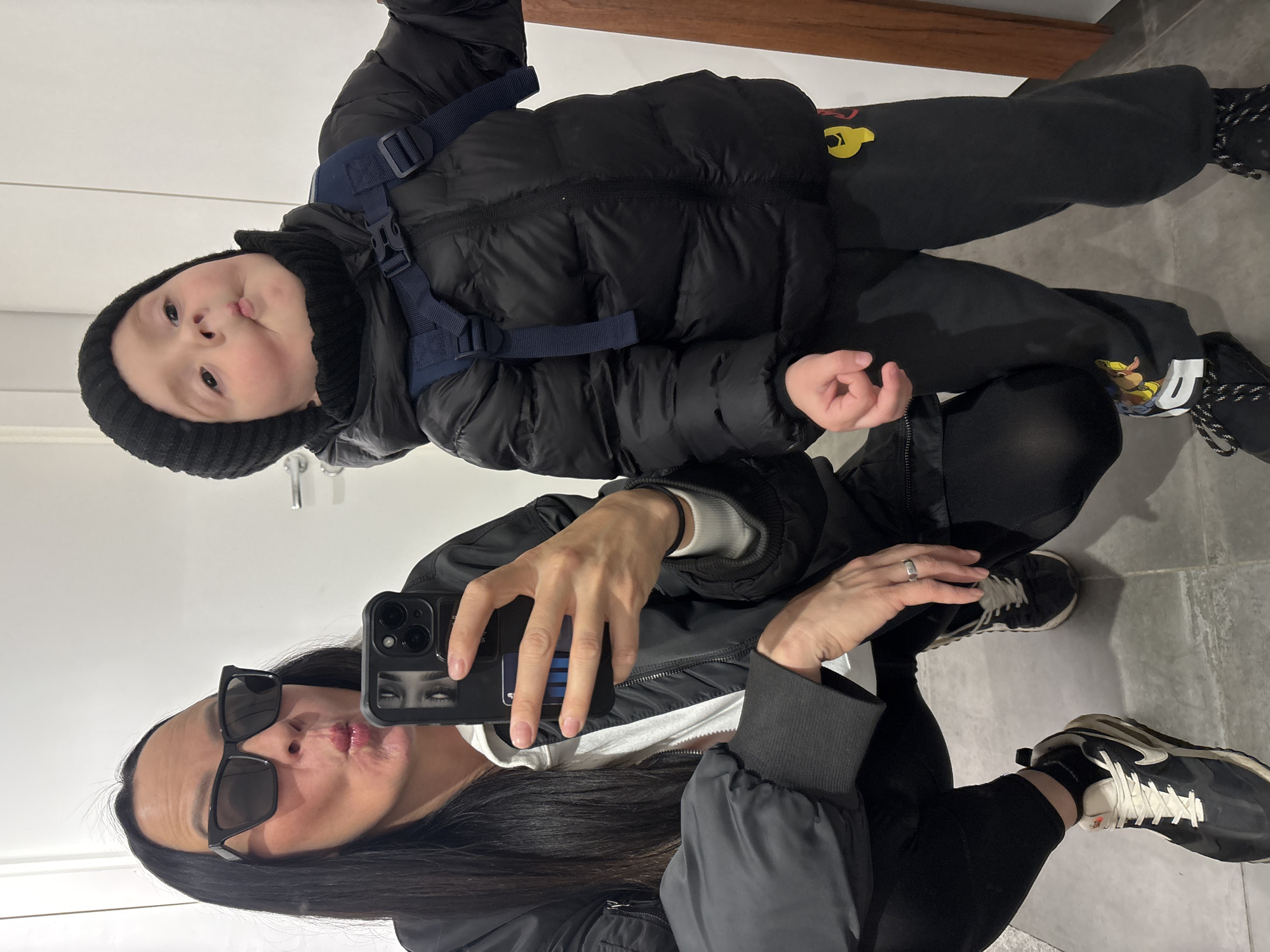 A woman takes a selfie in a mirror with a small child in Chaikovsky, Russia. The woman is wearing sunglasses and a black jacket and pants, while the child has on a black puffer jacket, black knit hat, black pants with yellow cartoon characters and black shoes. The room has tile flooring.