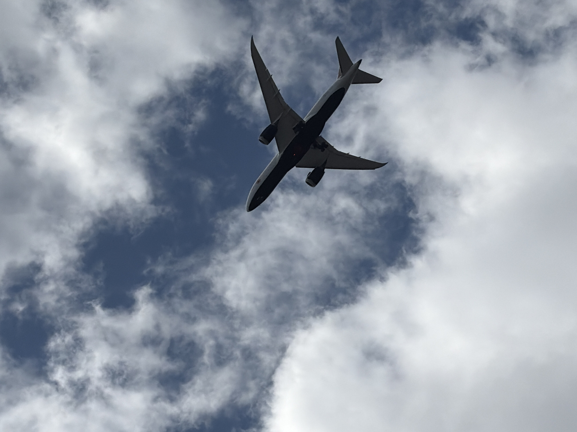 An airplane is captured from below, ascending into a partly cloudy sky. The aircraft appears to be a wide-body jet with two engines mounted under its wings. The underside of the plane is dark, contrasting with the white sections of the fuselage and wings. The sky is a mix of deep blue and white clouds, with the clouds casting some shadows on the upper surfaces of the airplane. The image was taken from the ground, looking directly upwards.