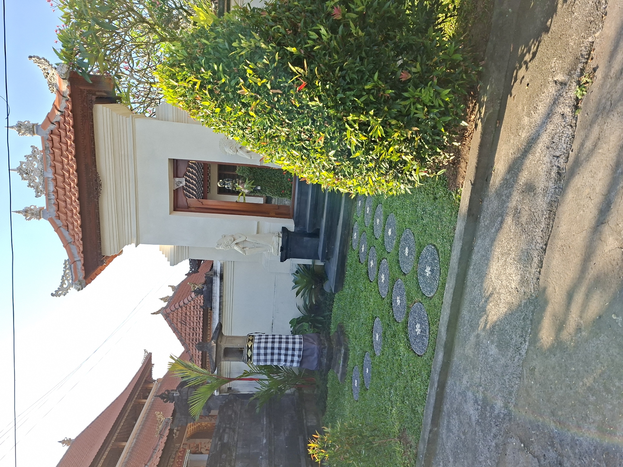 The image depicts the entrance to a building in Blahbatuh, Indonesia, with traditional Balinese architectural elements. A pathway made of circular stone pavers is laid across a green lawn. To the left, a stone lantern is adorned with a black and white checkered cloth. In front of the entrance, two white statues stand on pedestals. A large, lush green shrub occupies the right side of the frame. The building features a red-tiled roof with ornate decorative elements. The sky is clear and blue. The scene appears to be a residential or temple entrance, bathed in sunlight.