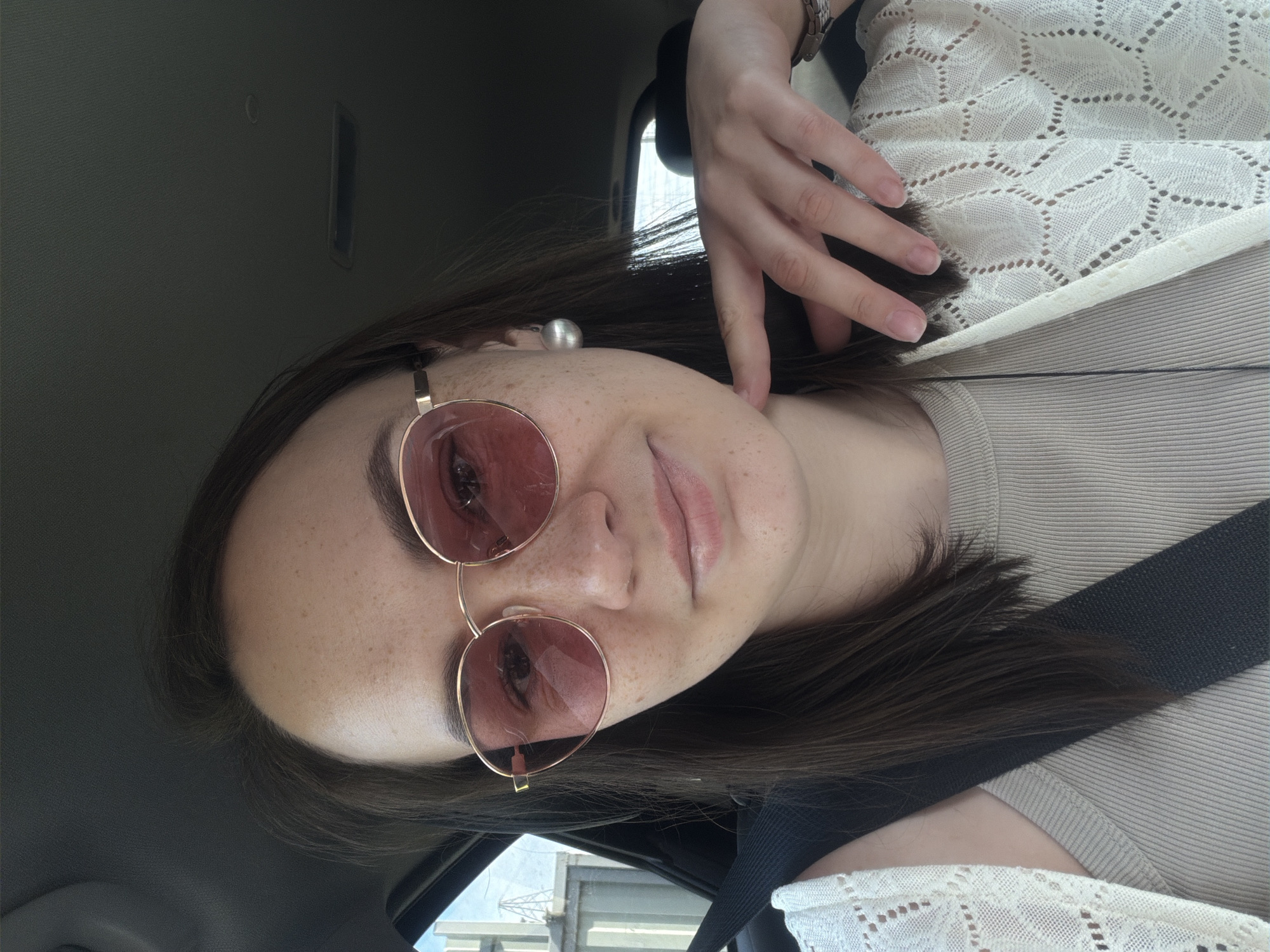 A young woman is in a car, wearing rose-tinted sunglasses and a white crocheted cardigan over a neutral-toned top. She has dark hair, freckles, and a pearl earring. Her hand is near her face. A seatbelt is visible across her chest. The interior of the car is visible, and outside the window, what appears to be a building and sky can be seen. The image gives no indication of whether it was taken in Chaikovsky, Russia.