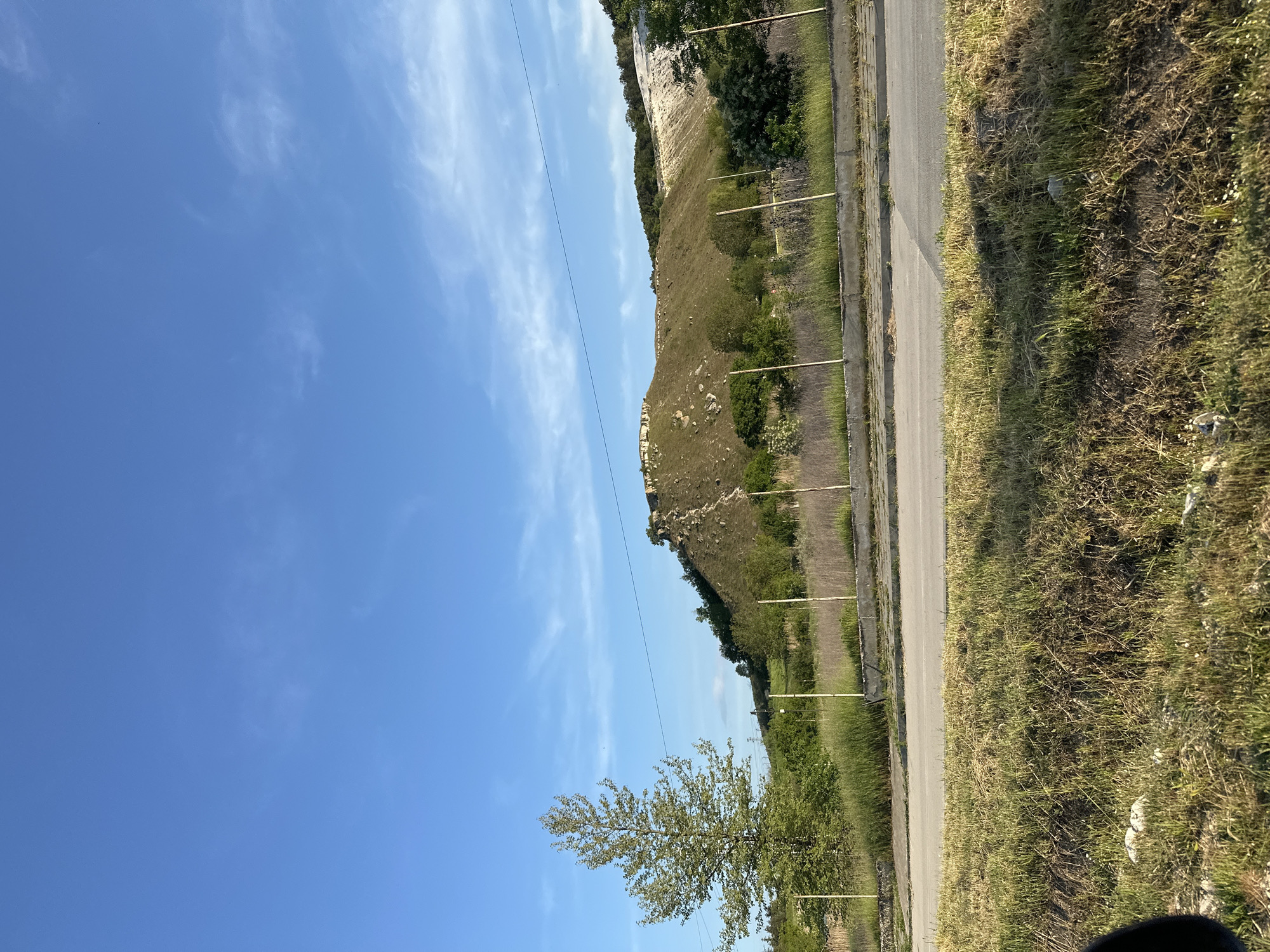 The image depicts a landscape in Beloslav, Bulgaria. A road runs along the right side of the frame, bordered by a concrete barrier and grassy verge. Parallel to the road is a hillside covered in vegetation. The top of the hill shows a cliff edge. A single tree is in the lower left of the image, while power lines extend across the sky. The sky is clear blue with some wispy white clouds.