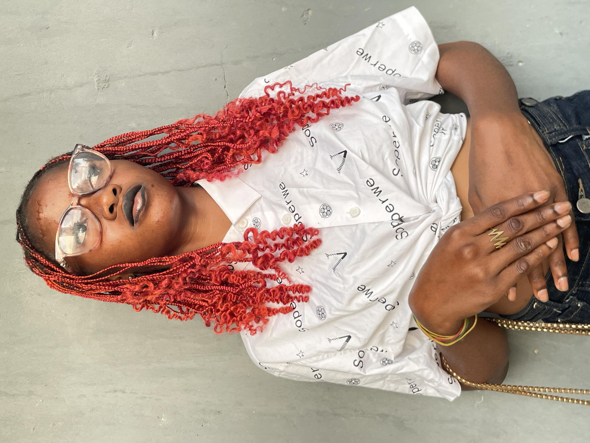 This medium shot captures a person lying down outdoors in Badagry, Nigeria, with their head positioned towards the left of the frame. The subject has dark skin, is wearing clear-framed eyeglasses, and has dark lipstick. Their hair is styled in vibrant red braids, with some sections featuring tight, bouncy curls.
They are dressed in a white short-sleeved button-up shirt, which is covered with black text and various symbols. The visible text on the shirt repeatedly features the word "Soperwe" and fragments like "per," "er," "So," "os," and "cepere." Symbols include stars, a geometric triangle-like shape ("A"), and a circular emblem resembling a stylized flower or gear. The person is also wearing dark blue jeans.
Accessories include multiple rings on their right hand, notably a distinctive gold-colored zigzag ring. On their left wrist, several thin, multi-colored bracelets in red, yellow, and green are visible. A gold-colored chain or strap is also seen near their lower body. The person appears to be in a relaxed pose, possibly looking directly at the camera. The background is a plain, light gray concrete surface, suggesting they are lying on a paved area or floor under daylight conditions.