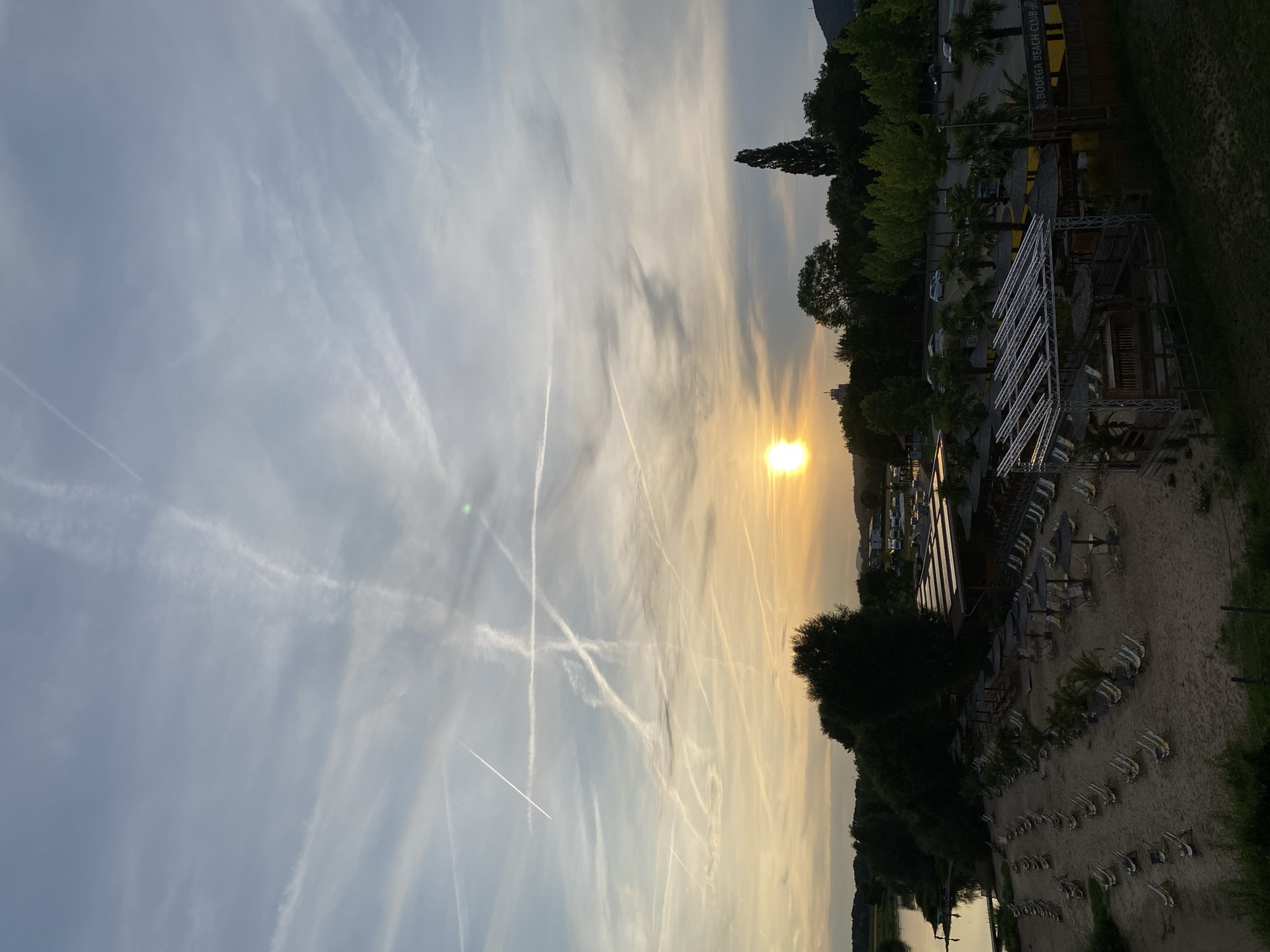 This media file captures a sunset scene at a beach club in Rinteln, Germany. The sun is low on the horizon, casting a warm, golden glow across the sky, which is filled with numerous contrails from aircraft, creating a dramatic, streaky pattern.
The foreground features a sandy beach lined with rows of empty lounge chairs and umbrellas, suggesting a peaceful, post-activity atmosphere. A structure resembling a beach bar or club building is visible, with a modern metal framework supporting a roof, and some wooden elements. Palm trees are interspersed among the seating, adding to the beach club ambiance. In the background, lush green trees line the edge of the property, and a body of water, possibly a river or lake, is visible. A mountain or hill can be seen in the far distance.
The overall mood is serene and tranquil, with the beautiful sunset and the deserted beach creating a sense of calm. The time of day is clearly sunset, and the weather appears clear with fair visibility, despite the abundance of contrails. Visible text on the building reads "BODEGA BEACH CLUB". No people are visible in the image.