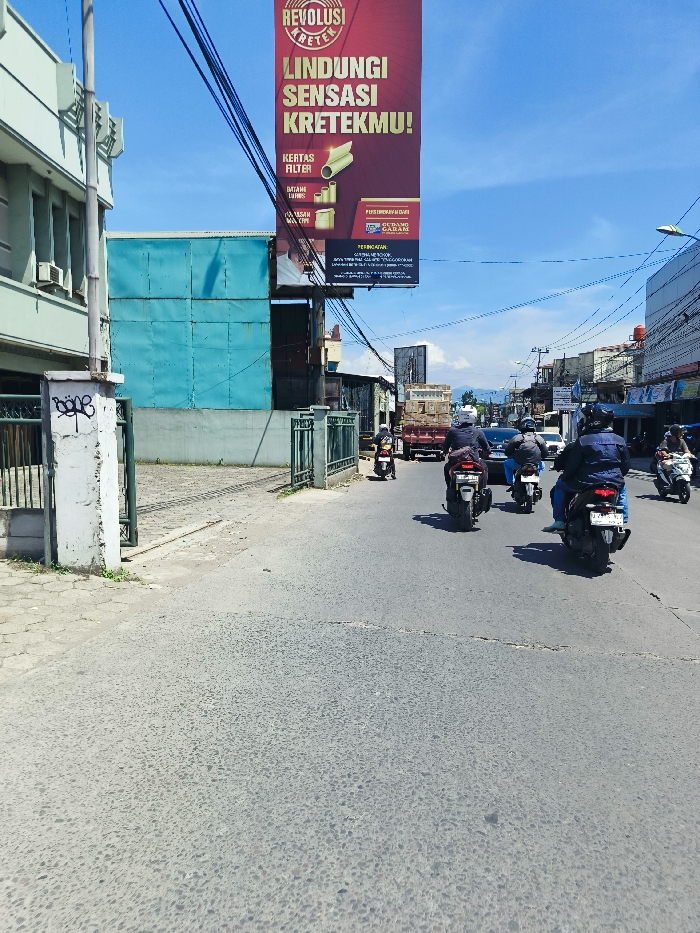 The image shows a street corner in Margahayu, Indonesia. It is a sunny day with a blue sky. A large red sign is mounted on a pole at the intersection. The sign reads, "Revolusu Kretrek, Lindungi Sensasi Kretrekmu!." On the street, several people are riding motorcycles. In the foreground, a white concrete pillar is painted with black graffiti that says "BODE". The street has a grey asphalt surface and the surrounding buildings are made of brick and concrete. There is a green metal fence to the left of the street. In the distance, additional buildings and trees are visible. The street appears to be a major road with a substantial amount of traffic. The traffic is mostly motorcycles but there are also some cars in the distance.