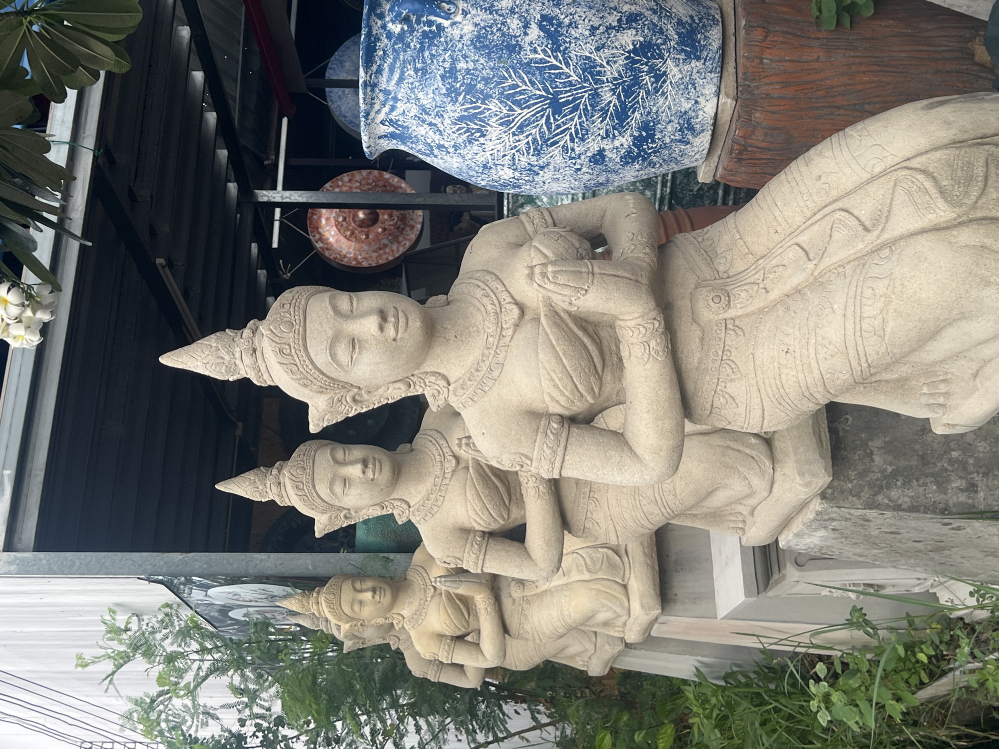 The image depicts a row of four identical stone statues of women with their hands pressed together in a prayer-like gesture. They are adorned with traditional headdresses and jewelry, and wear intricately carved clothing. The statues are arranged in a line, with the closest one in the foreground and the others receding into the background. To the right of the statues is a large, blue and white patterned ceramic vase. In the background, a corrugated metal structure suggests an outdoor market or stall. The scene is set against a backdrop of greenery and what appears to be a cloudy sky. The location is identified as Ko Samui District, Thailand.