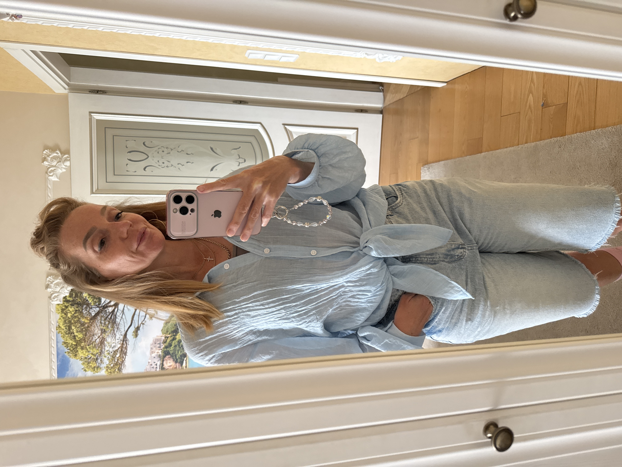 A woman is standing in front of a mirror in a room. She is wearing a light blue, crinkled button-down shirt tied at the waist, denim shorts, and pink socks. She is holding a pink cell phone with a keychain attached and has her left hand in her pocket. The room has light-colored walls, a wooden floor, and a landscape painting on the wall. The mirror reflects the woman and parts of the room.