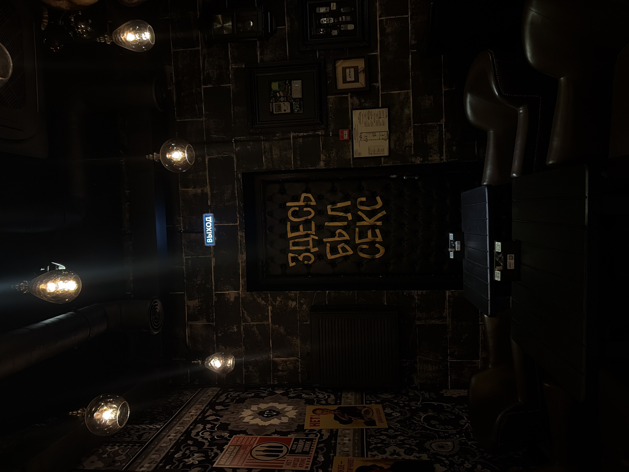 The image depicts the interior of a dimly lit bar in Saint Petersburg, Russia. The walls are constructed of dark, rough-hewn bricks, creating a rustic ambiance. On the left side, a patterned wallpaper features an ornate floral design in shades of grey and black. Several vintage posters with Russian text are visible on this wall.
The ceiling is adorned with industrial-style pendant lights, each housing a warm, glowing Edison bulb. A large ventilation duct runs horizontally across the ceiling. A bright blue "EXIT" sign is illuminated above a prominent doorway in the center of the frame. This door is upholstered with dark material, and large, yellow, hand-painted Cyrillic letters spell out "ЗДЕСЬ БЫЛ СЕКС" which translates to "SEX WAS HERE".
To the right of the door, the brick wall is decorated with several framed items. These include what appear to be collages of old video game consoles and cartridges, and a framed cassette tape. Further down, there is a framed display of vintage mobile phones. A small red fire alarm is also mounted on the wall.
In the foreground, two dark, rectangular tables are positioned, with their surfaces reflecting minimal light. Comfortable, dark brown leather armchairs are placed around the tables, some of which are partially obscured by shadow. The overall atmosphere is intimate and slightly mysterious, with the lighting casting long shadows and highlighting the textures of the materials.