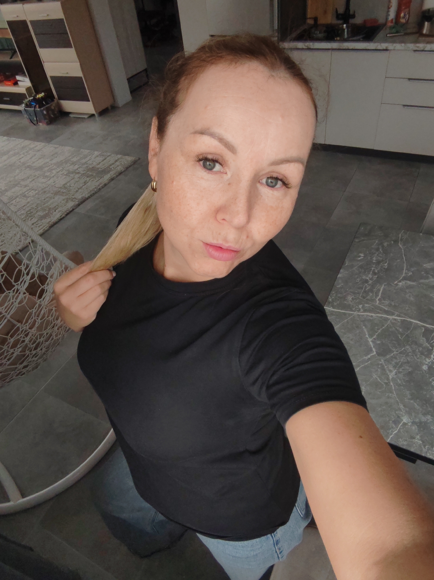 A fair-skinned woman with freckles is seen in a selfie-style shot. She has blonde hair, visible gold hoop earrings, and is wearing a black T-shirt and light blue jeans. The location appears to be indoors, with elements suggesting a domestic setting. In the background, there are light-colored kitchen cabinets and a dark gray countertop. Additionally, a decorative rug and what appears to be a hanging chair are partially visible. The image does not provide enough information to confirm the city of Chaikovsky, Russia.