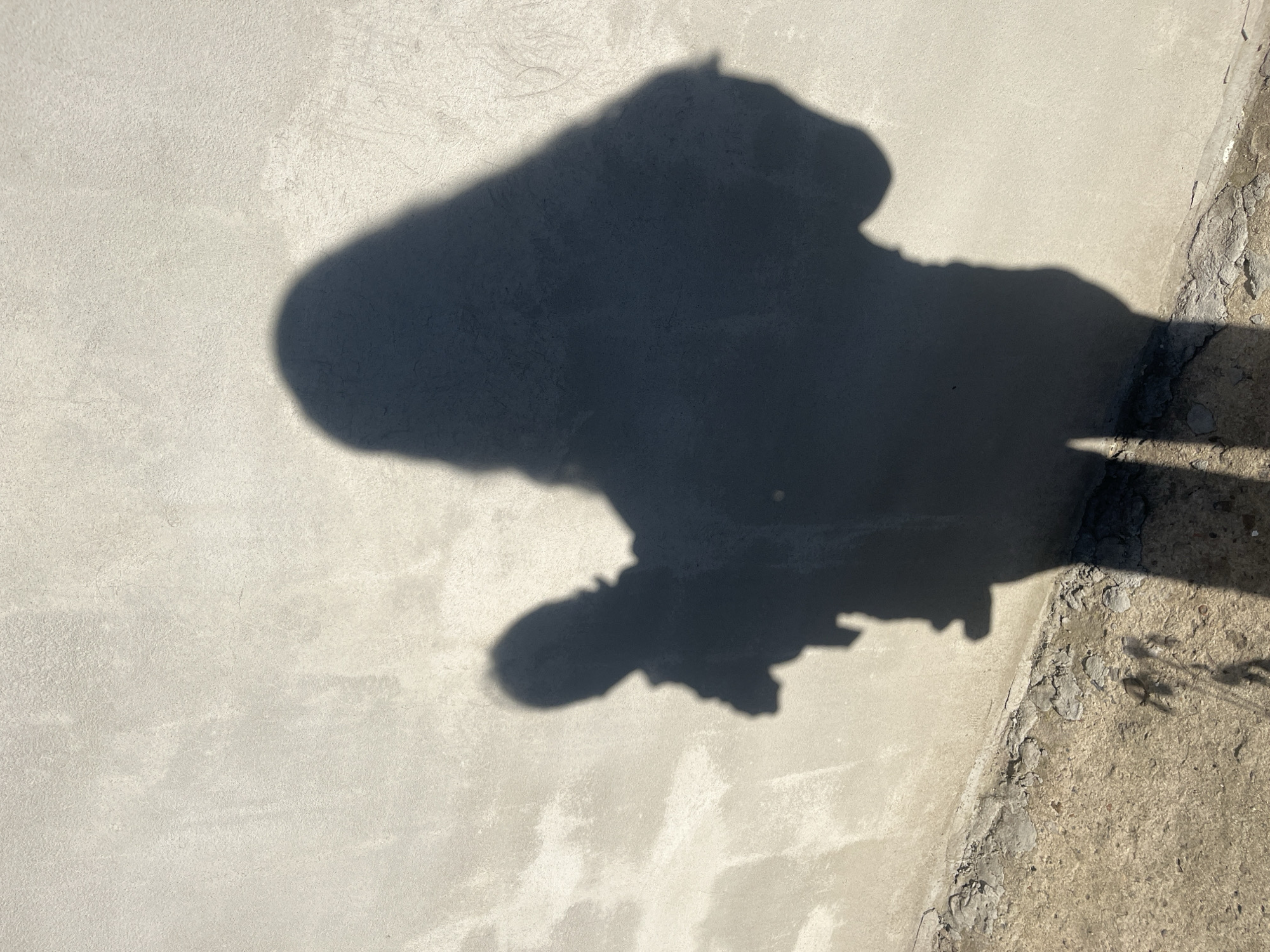 The image displays a shadow of a person on a textured, light-colored surface, likely concrete or stucco. The shadow is dark and sharply defined, suggesting bright, direct sunlight. The figure in the shadow appears to be wearing clothing, with a discernible head and torso, and possibly legs. The overall impression is that of a person standing or walking outdoors. The surface has a rough, granular texture with some variations in color and smoothness. In the lower right portion of the image, a transition to a rougher, uneven surface is visible, with cracks and debris, suggesting an outdoor environment. The light and shadow indicate it is likely daytime. No text is visible in the image.
