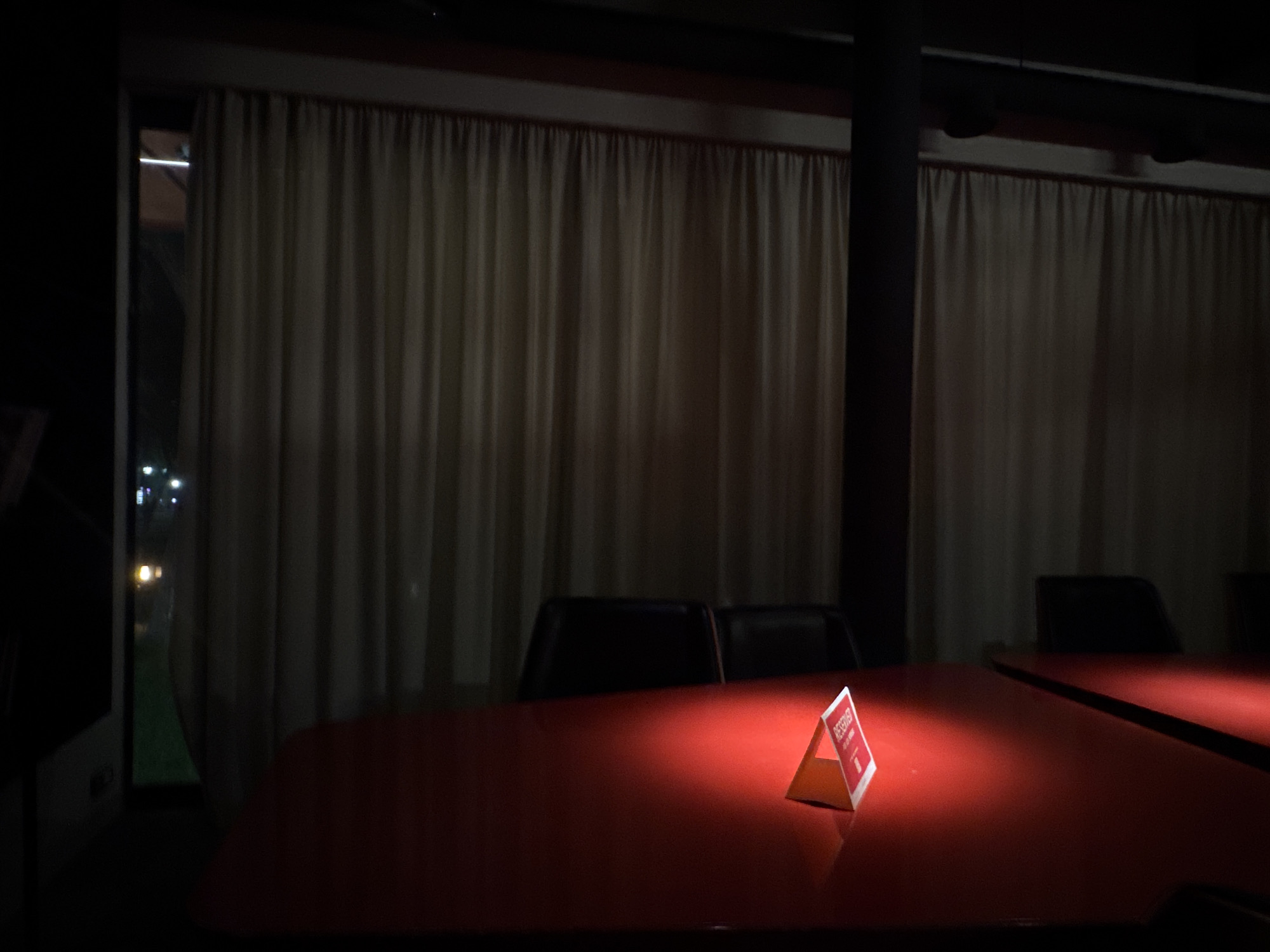 The image depicts an interior scene, likely a dining area or function room, potentially located in Yerevan, Armenia. The room appears dimly lit, with light sources not readily apparent but casting a spotlight on a red table in the foreground. On the table, a triangular "Reserved" sign stands out. Behind the table, there are chairs positioned facing the camera. Heavy, light-colored curtains are draped in the background, obscuring the view outside, though a glimpse of an exterior scene is visible through a window on the left, showing blurred lights suggesting a cityscape at night. A dark, vertical structural support is also present in the background, situated between the curtains.
