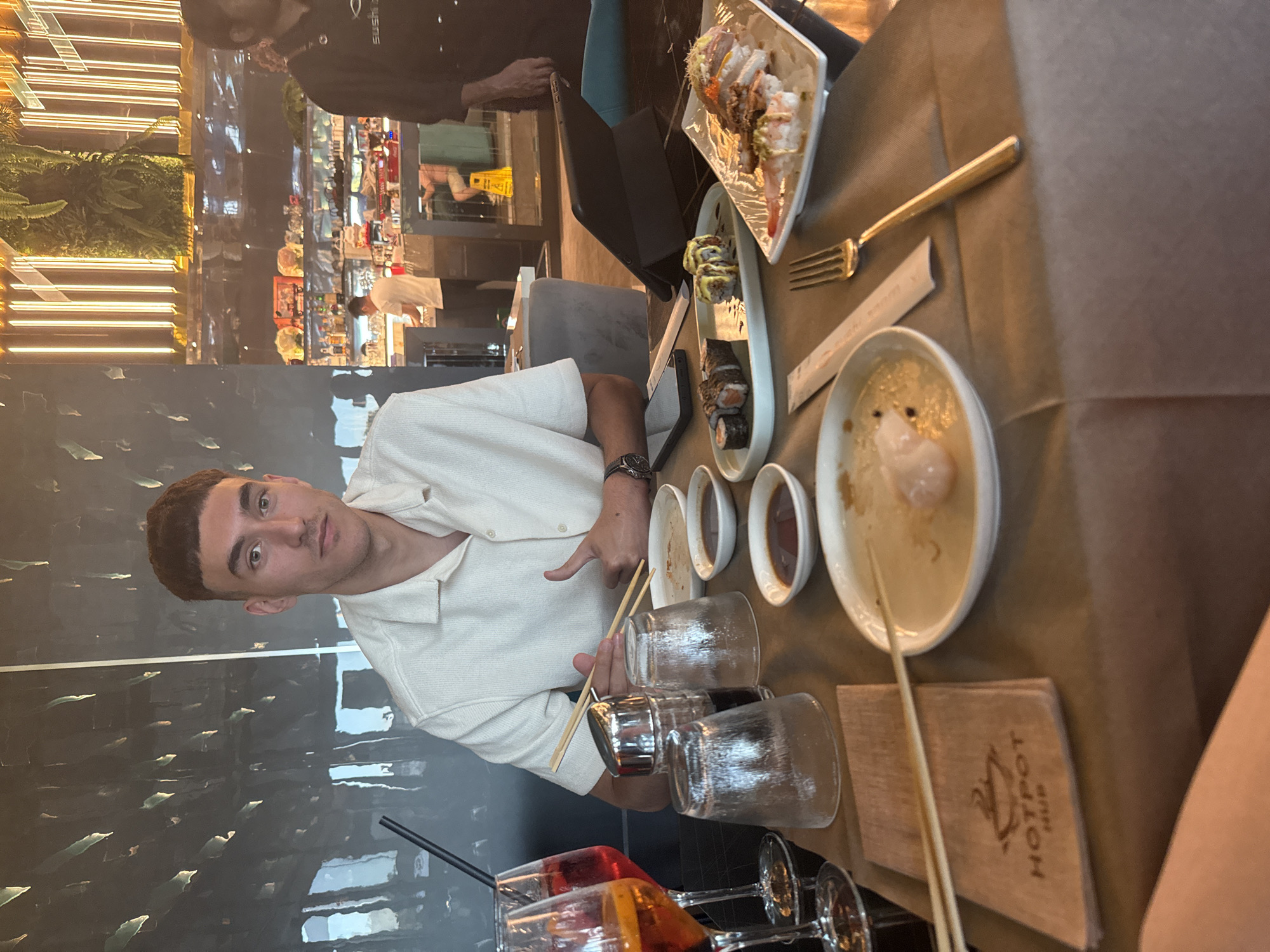 A medium shot, rotated 90 degrees counter-clockwise, shows a young man seated at a table in what appears to be a modern restaurant or bar. The man has short, dark hair and is wearing a light-colored, collared shirt and a dark watch on his left wrist. He looks directly at the camera with a neutral expression, holding chopsticks in his left hand and making a pointing gesture with his right hand.
The table in front of him is covered with a dark brown placemat or tablecloth. Several dishes are present, including plates of sushi rolls and what looks like a partially eaten dessert in a white bowl. Small bowls of dark sauces (likely soy sauce) are also visible. There are multiple empty clear glasses and two glasses containing reddish-orange drinks with straws, suggesting cocktails. A golden fork rests on the table, along with a rectangular placemat or menu cover that reads "HOTPOT HUB".
The background features a contemporary interior with warm, horizontal strip lighting on a wall adorned with greenery. A dark, reflective wall is behind the man. Other patrons and staff are visible in the blurred background, suggesting a busy dining environment. A person in the far background wears a black top with "SUSHISU" visible on it. The overall setting suggests an evening meal or social gathering at an Asian-themed or fusion restaurant.